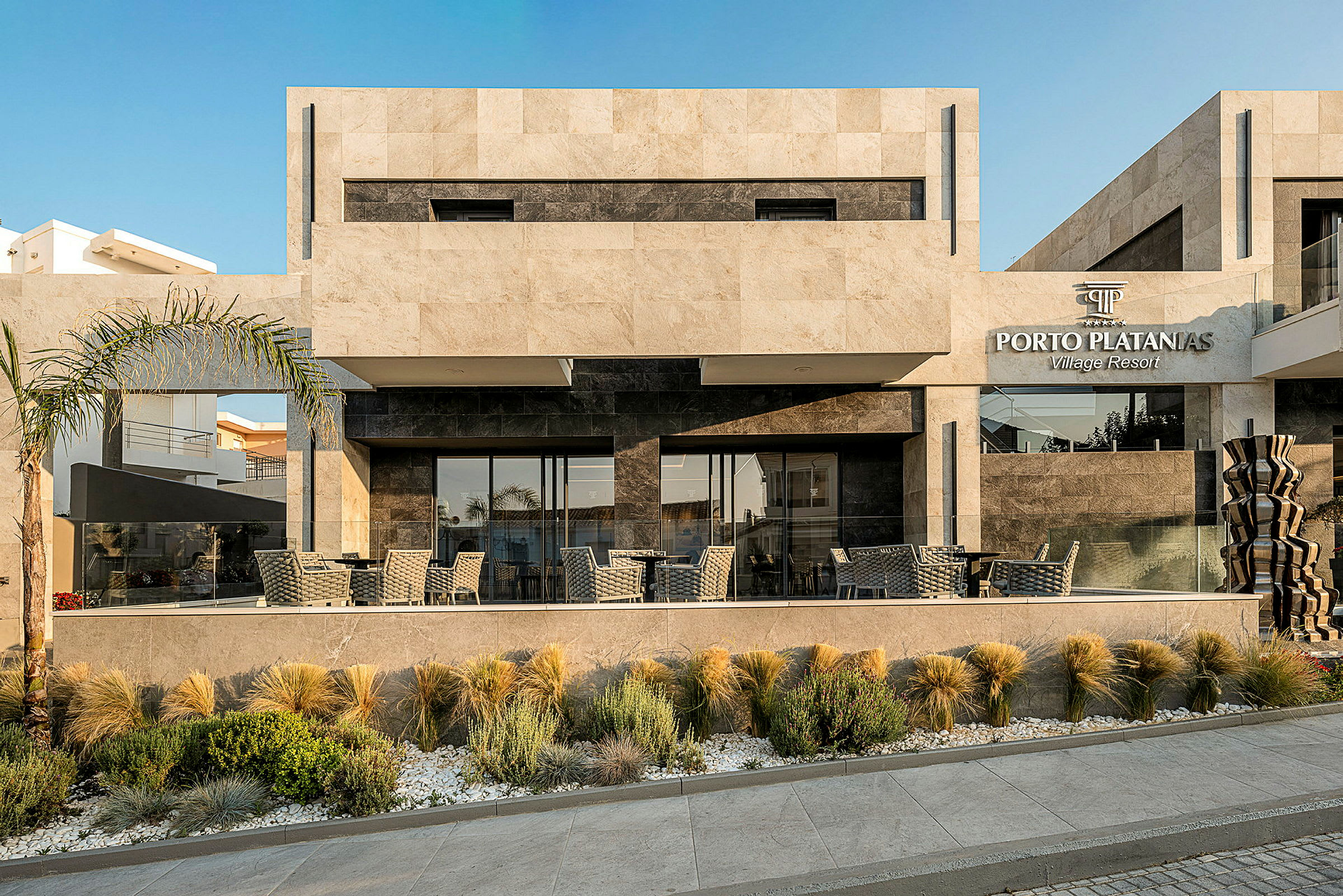 Exteriör av Porto Platanias Village Resort byggnad med modernt design och uteservering. (AI-genererad text)