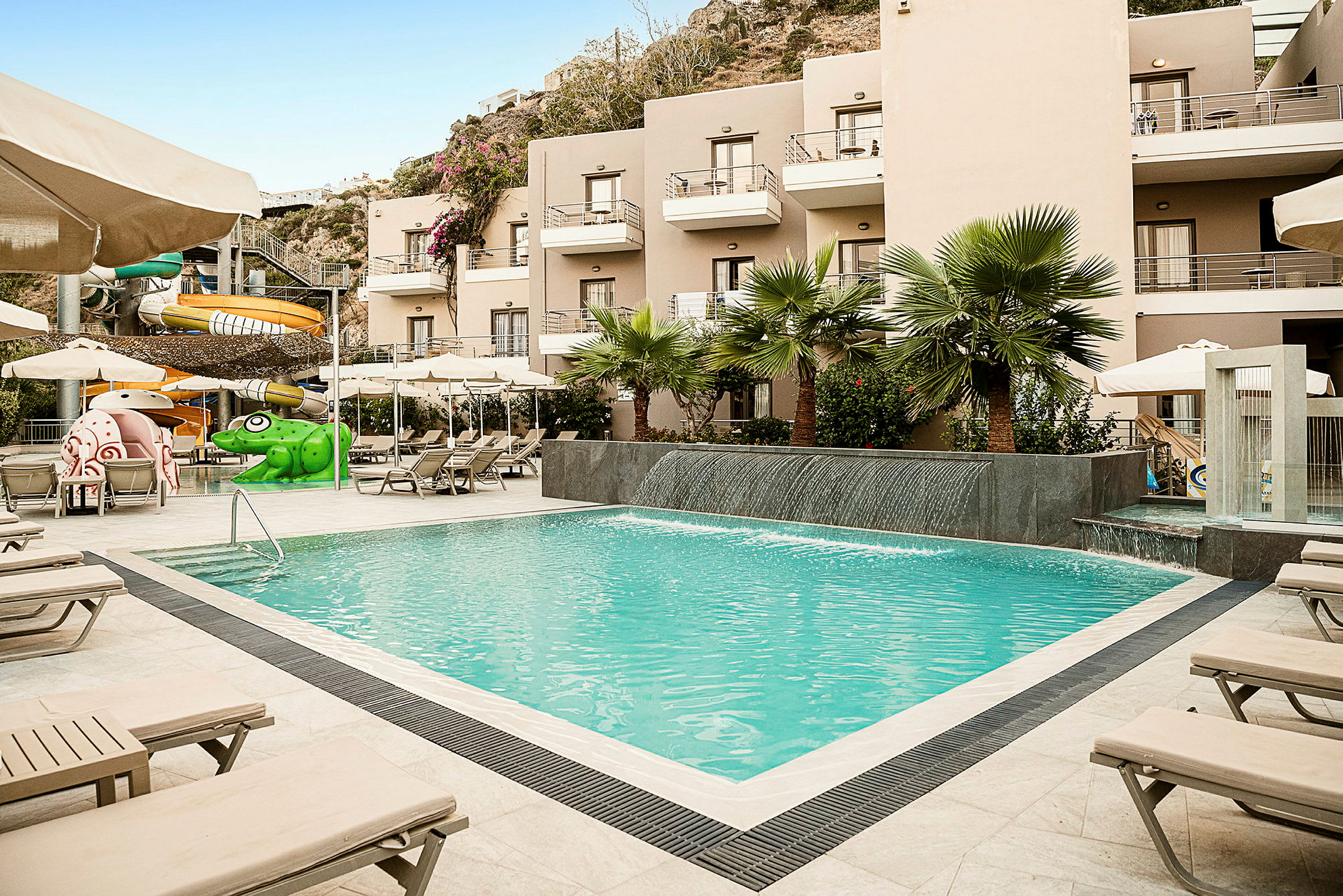 En utomhuspool bredvid ett hotell med solstolar, vattenrutschbanor och grönska. (AI-genererad text)
