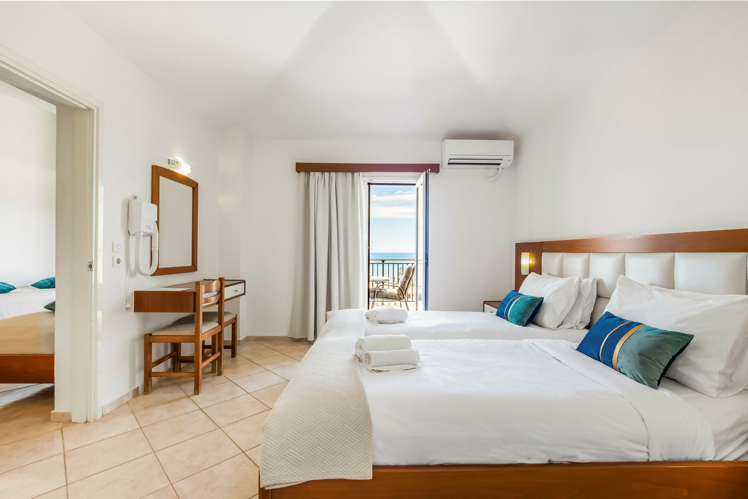 Ett hotellrum med två enkelsängar, sänglampor och en sittgrupp vid balkongen som har utsikt mot havet. (AI-genererad text)