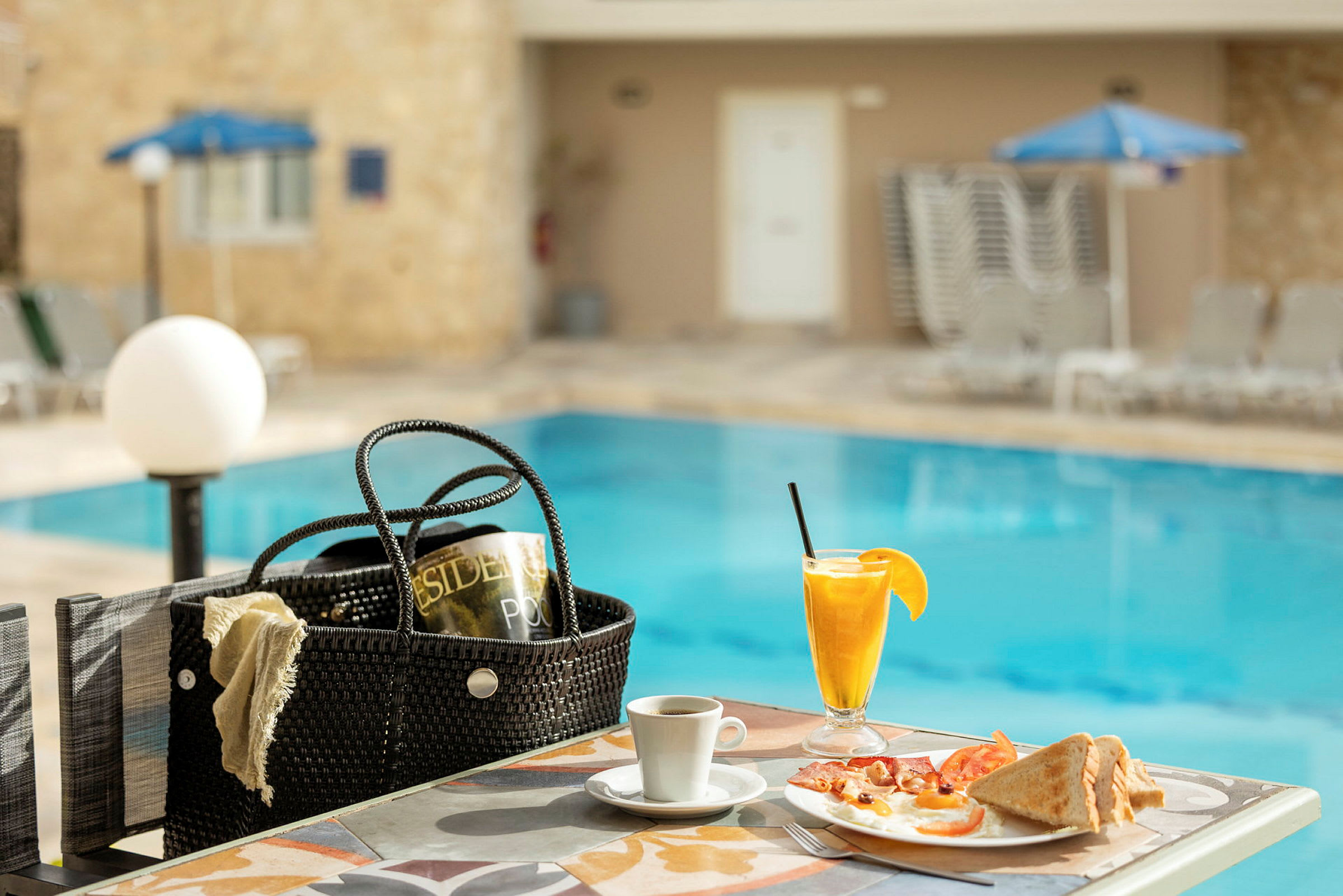 Frukost serverad vid ett bord nära en swimmingpool, inklusive apelsinjuice, kaffe och en tallrik med bröd och ägg. (AI-genererad text)