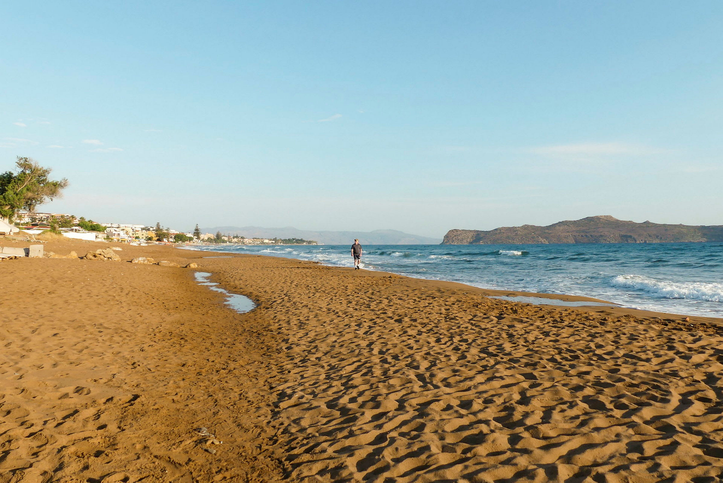 En sandstrand vid havet med en ensam person som promenerar nära vattenkanten. (AI-genererad text)