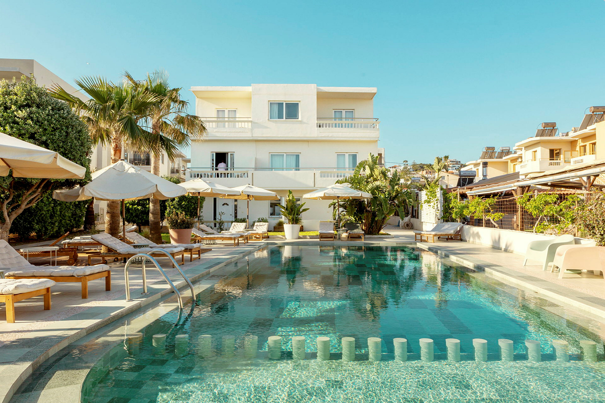 Hotellbyggnad med en pool framför och omgivande solstolar samt parasoller. (AI-genererad text)