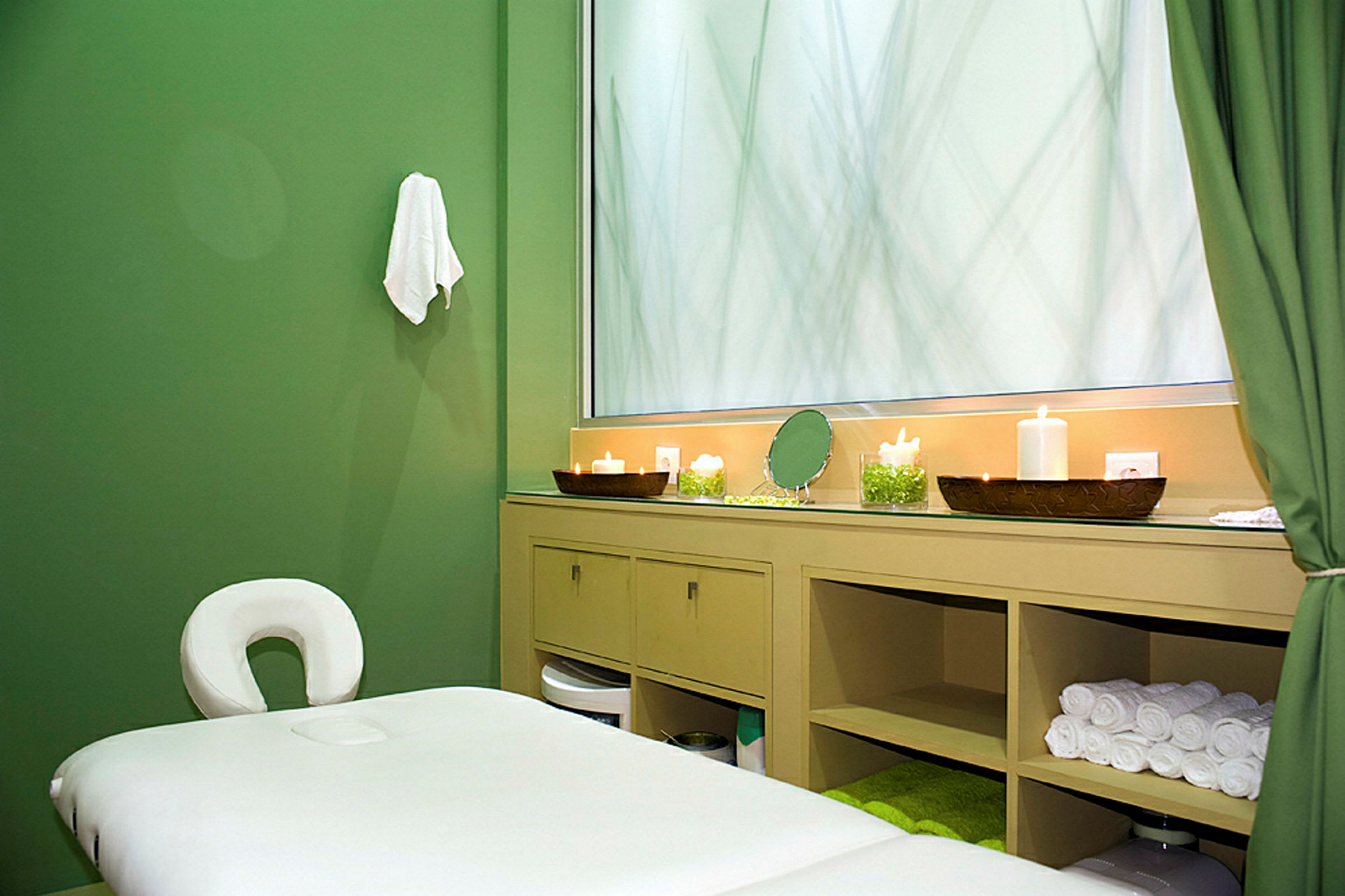 Ett massagebord i ett rum med gröna väggar och ett skåp med handdukar och ljus. (AI-genererad text)
