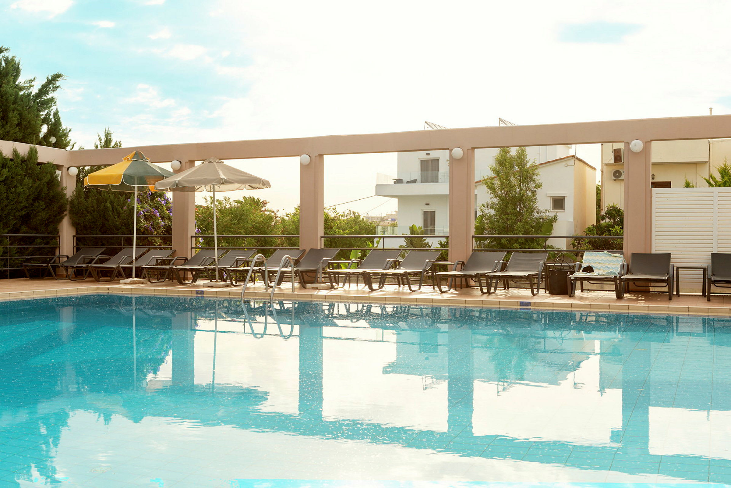 En pool med solstolar och parasoller runt kanten, omgiven av vegetation och byggnader. (AI-genererad text)
