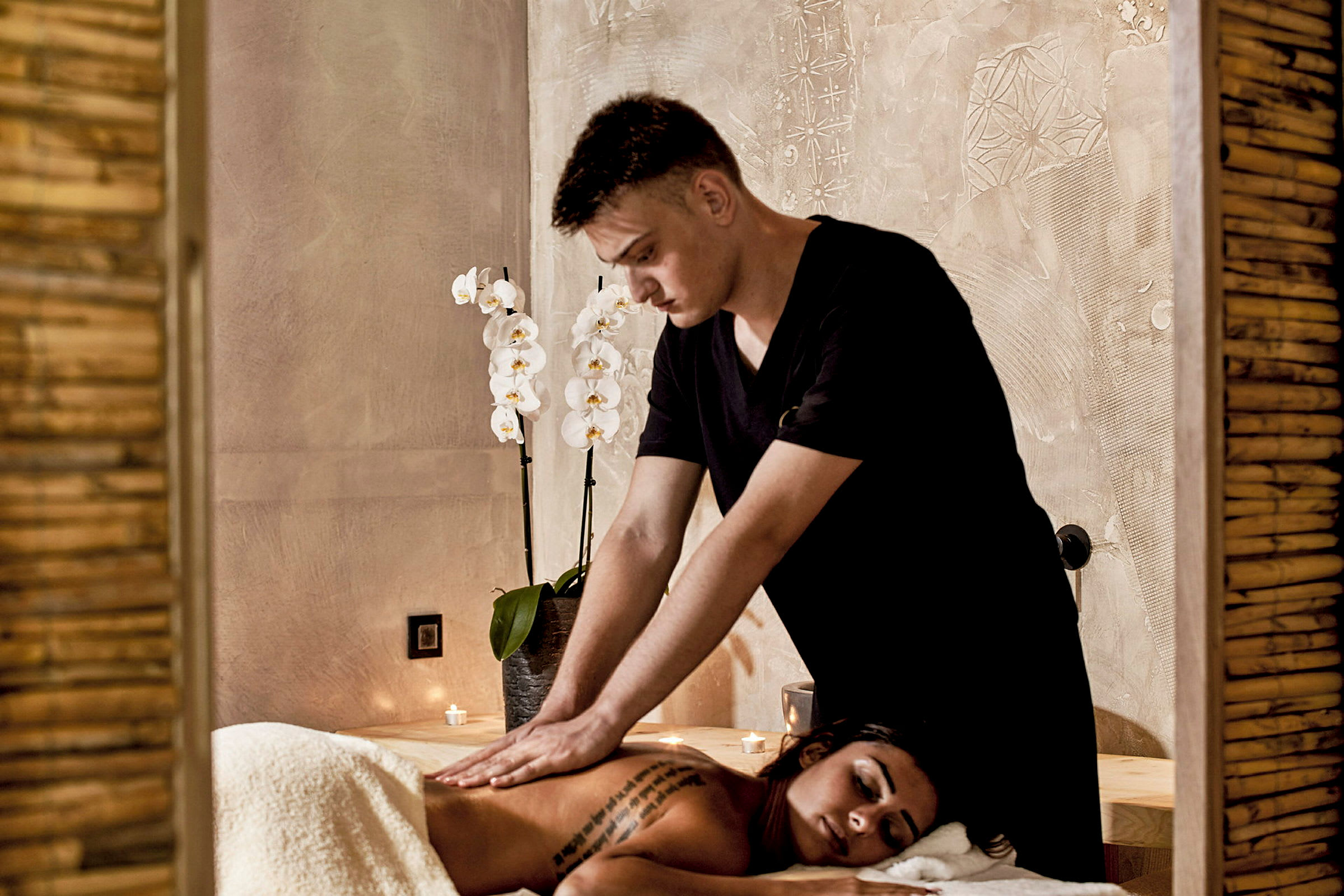 En person får en ryggmassage i ett rum med dekorativa vita orkidéer och levande ljus. (AI-genererad text)