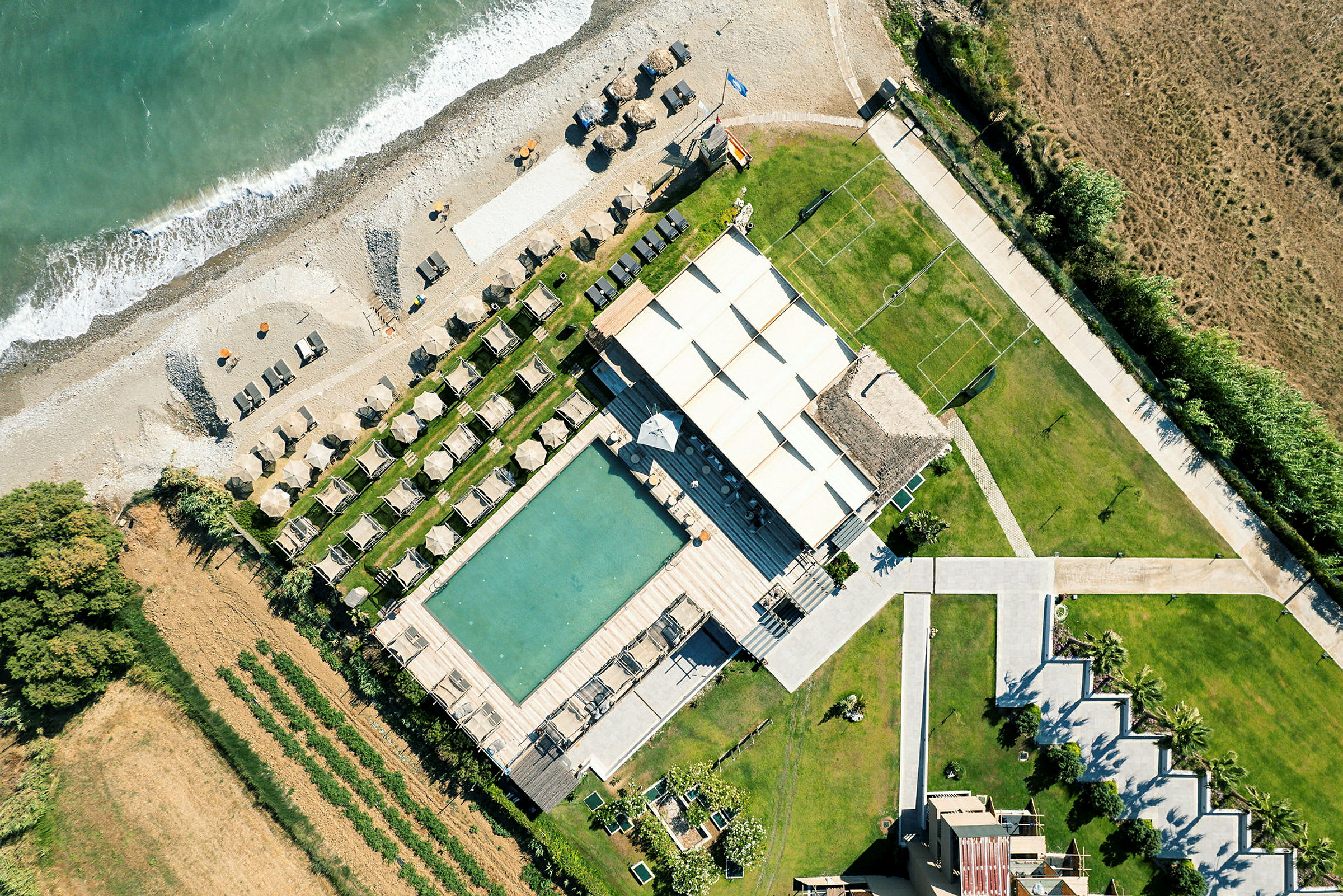 Flygfoto av en strandnära resort med solstolar, parasoller, pool och en omgivande grönskande miljö. (AI-genererad text)