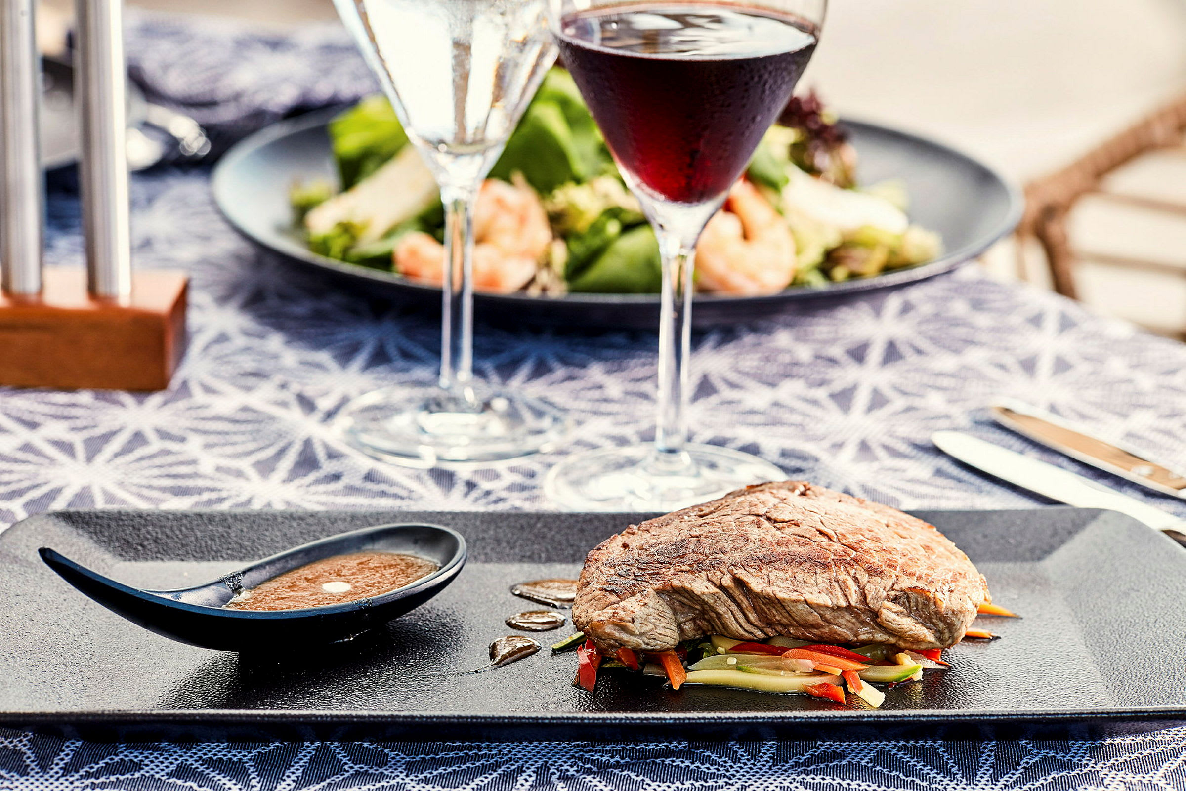 En tallrik med kött på grönsaker och en liten såsskål, med vinglas och sallad i bakgrunden. (AI-genererad text)
