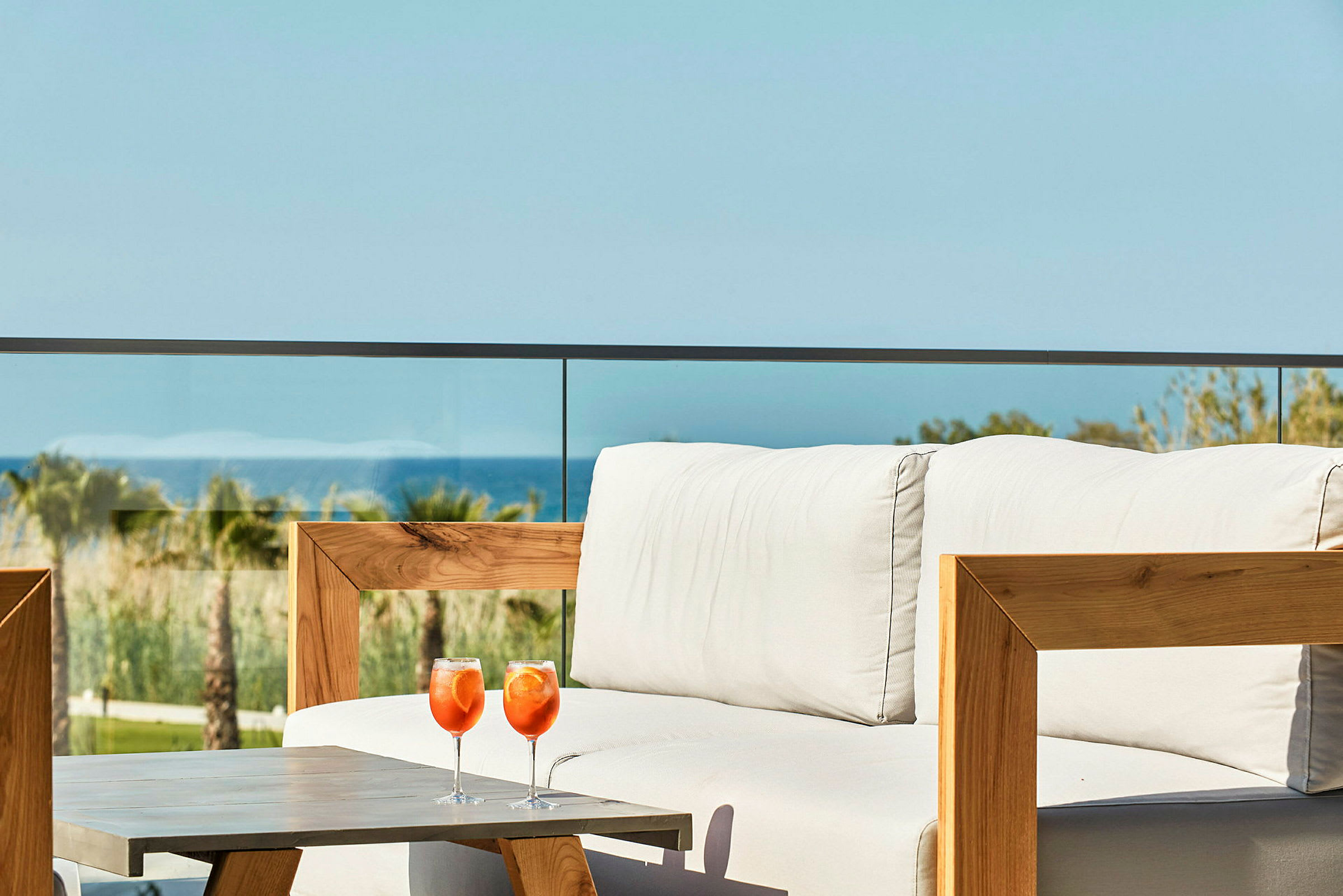 En soffa med vit klädsel och två orange drycker på ett bord framför, med utsikt mot havet och palmar i bakgrunden. (AI-genererad text)