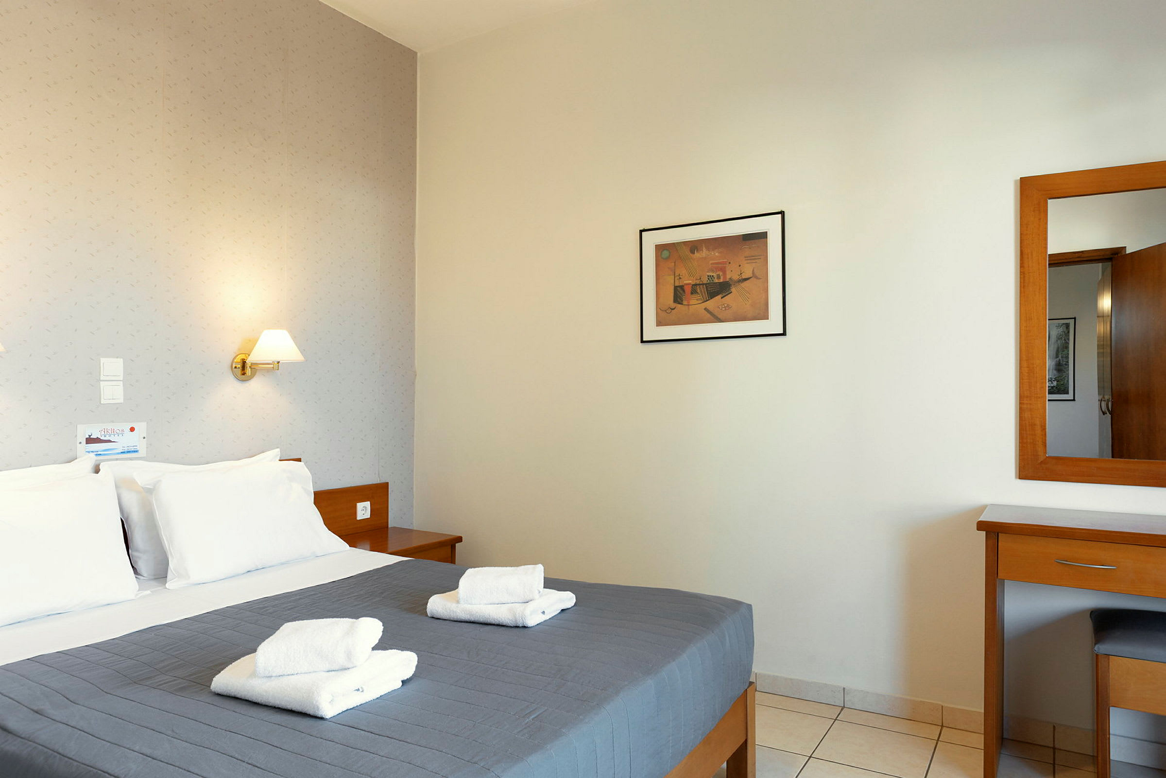 Ett hotellrum med en dubbelsäng och grått överkast. På sängen ligger två vikta vita handdukar. På väggen hänger en tavla och bredvid finns en spegel ovanför ett skrivbord. (AI-genererad text)