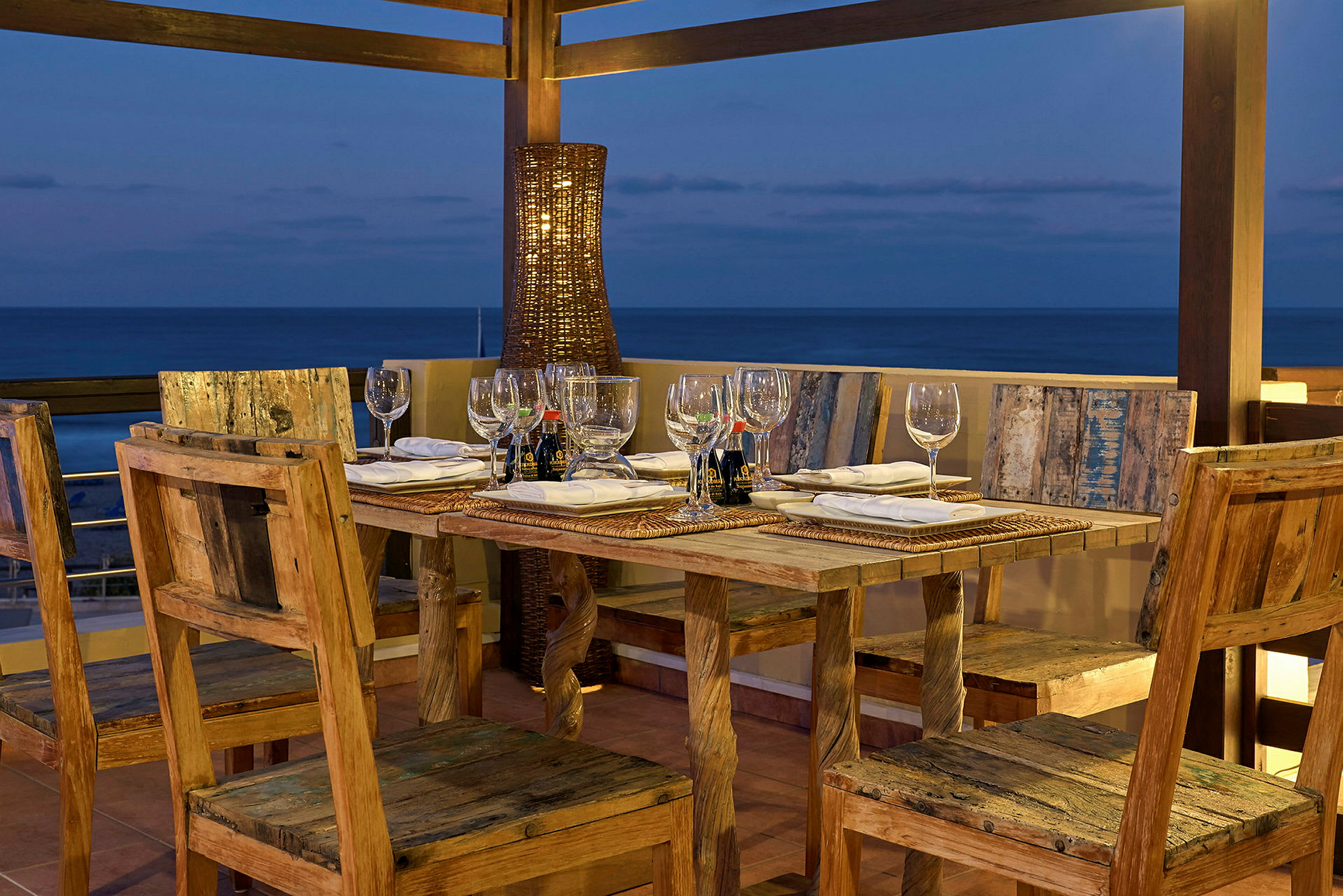 Ett rustikt matbord med trästolar är dukat med glas, servetter och tallrikar på en terrass med utsikt över havet i skymningen. (AI-genererad text)