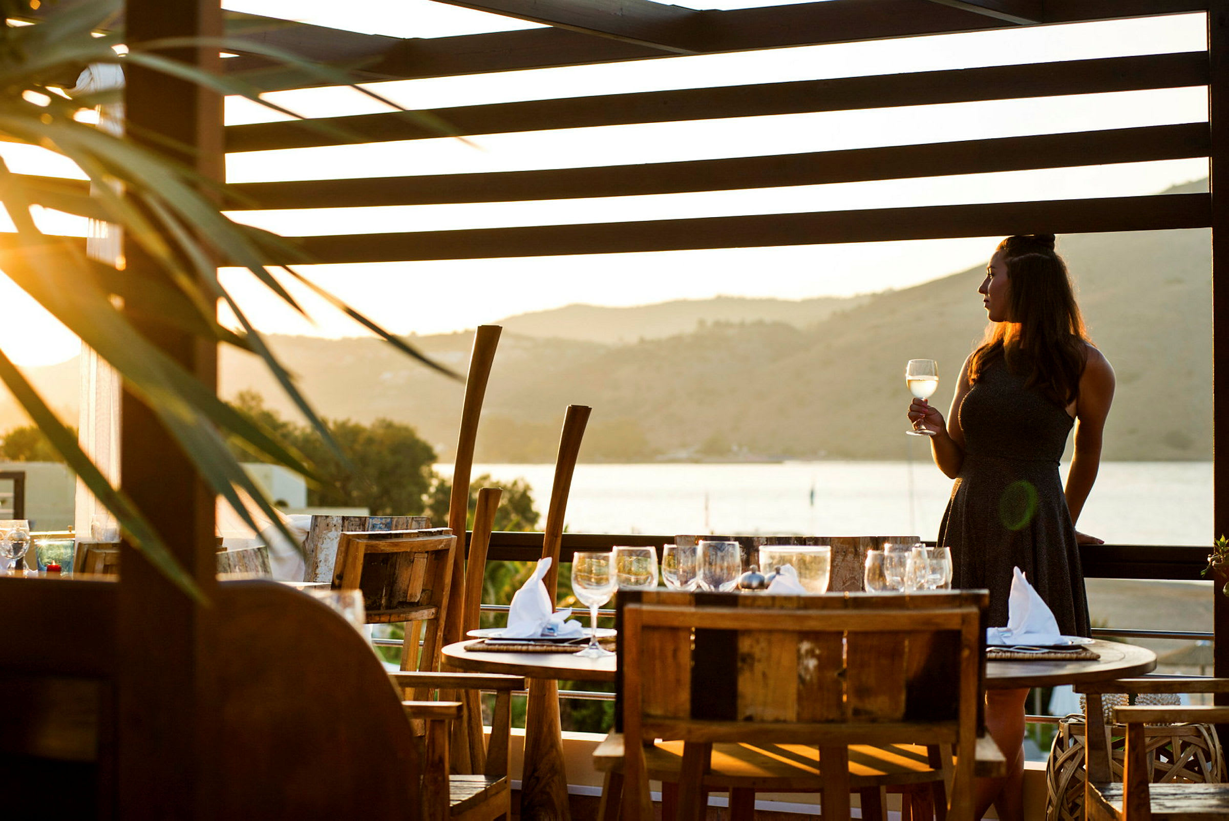 Person standing on a terrace with tables and chairs, holding a glass of wine, with a scenic view of a body of water and hills during sunset. (AI-genererad text)