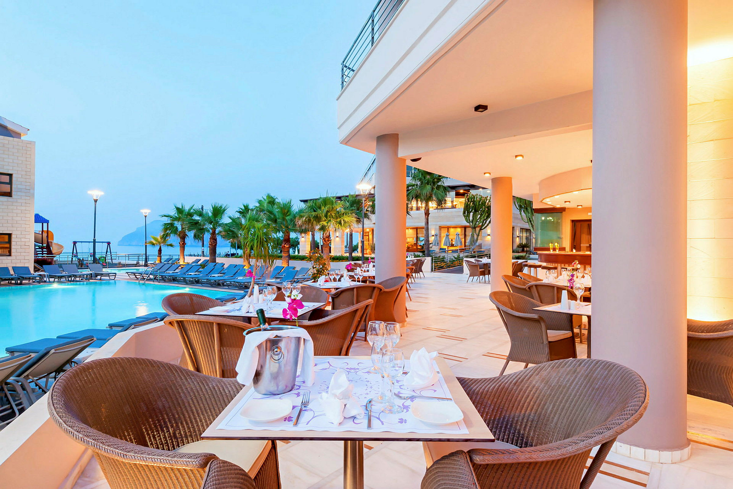 Restaurangområde vid en pool med bord och stolar, omgiven av palmer. (AI-genererad text)