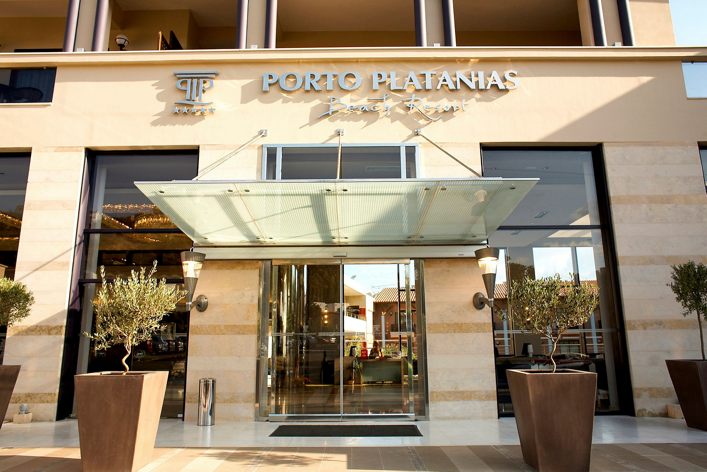 Entrén till Porto Platanias Beach Resort, med glasdörrar och planterade olivträd framför. (AI-genererad text)