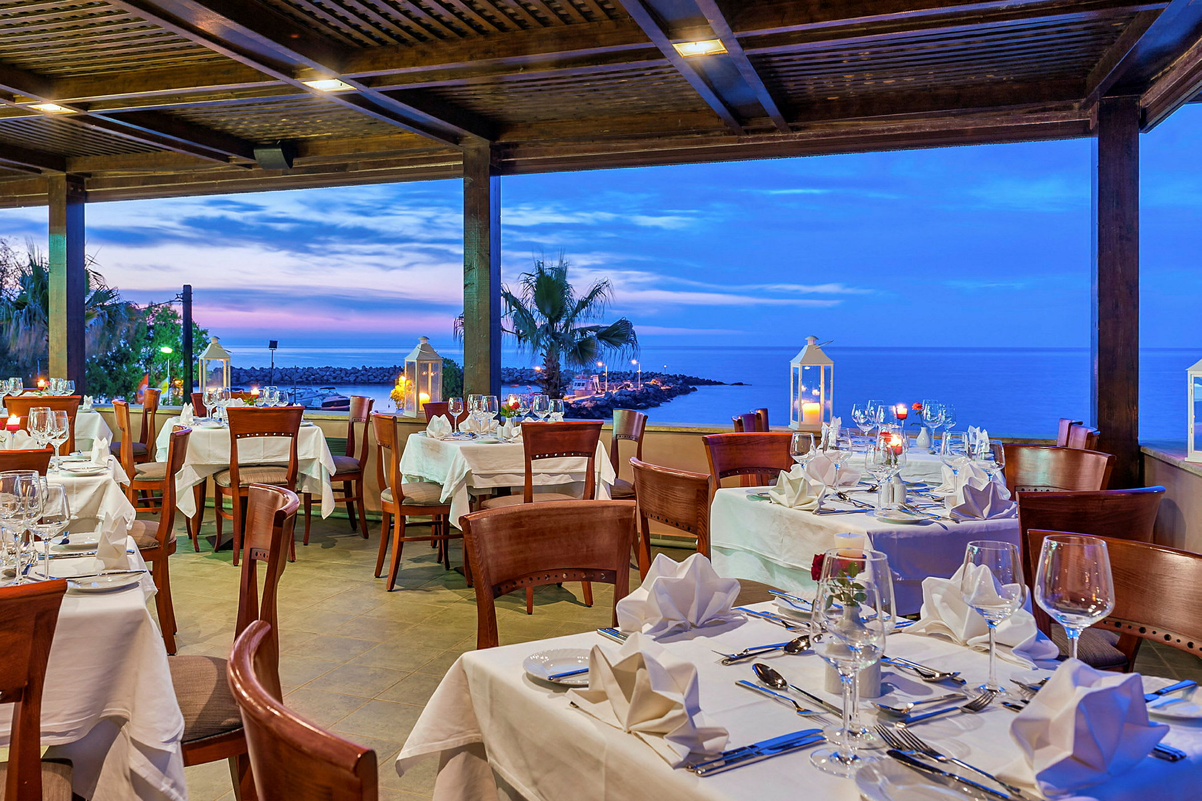 En restaurang med uppdukade bord med vita dukar, med utsikt över havet och solnedgången. (AI-genererad text)