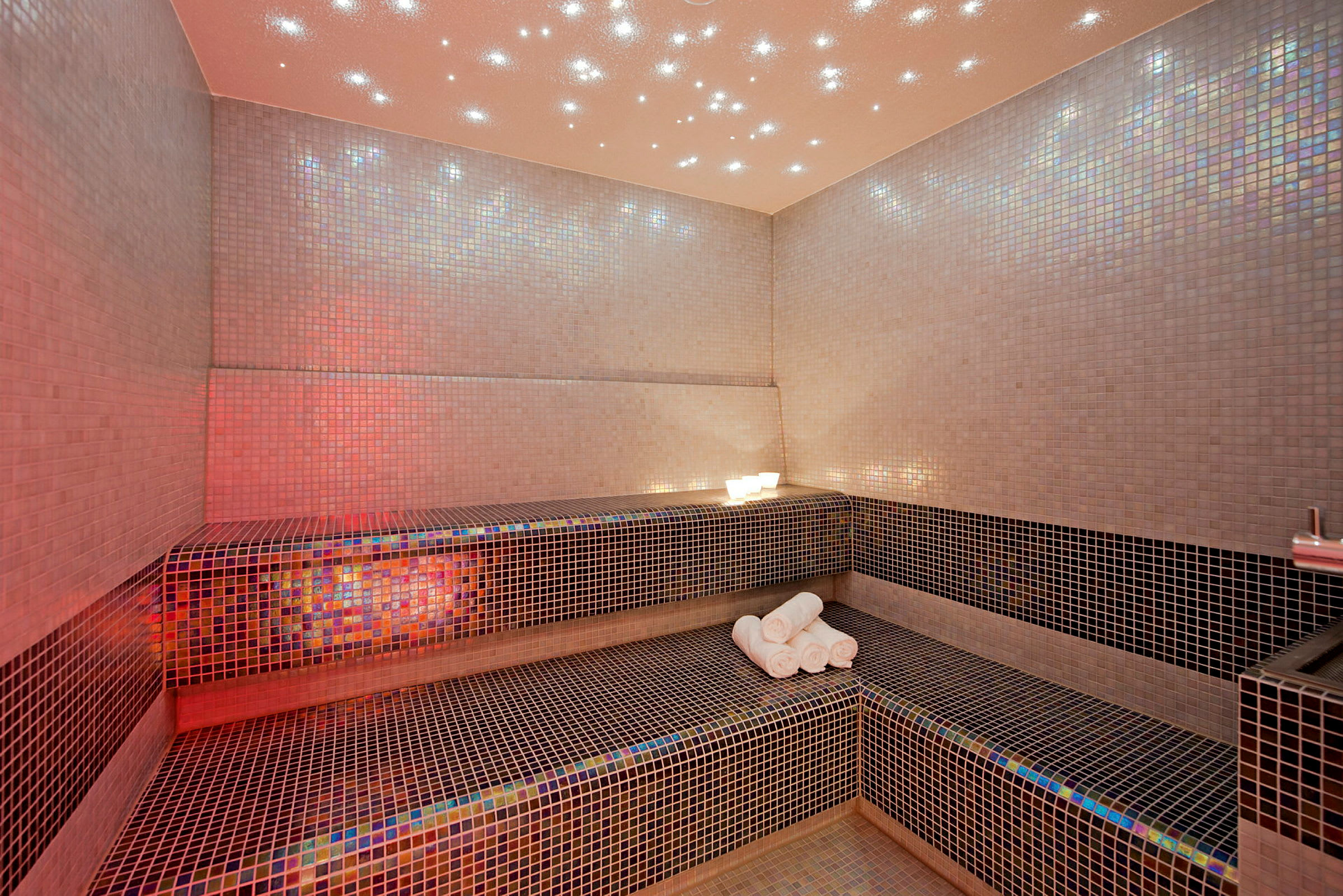 Rummet är ett mosaikkakelklätt spa med en bänk och handdukar. (AI-genererad text)