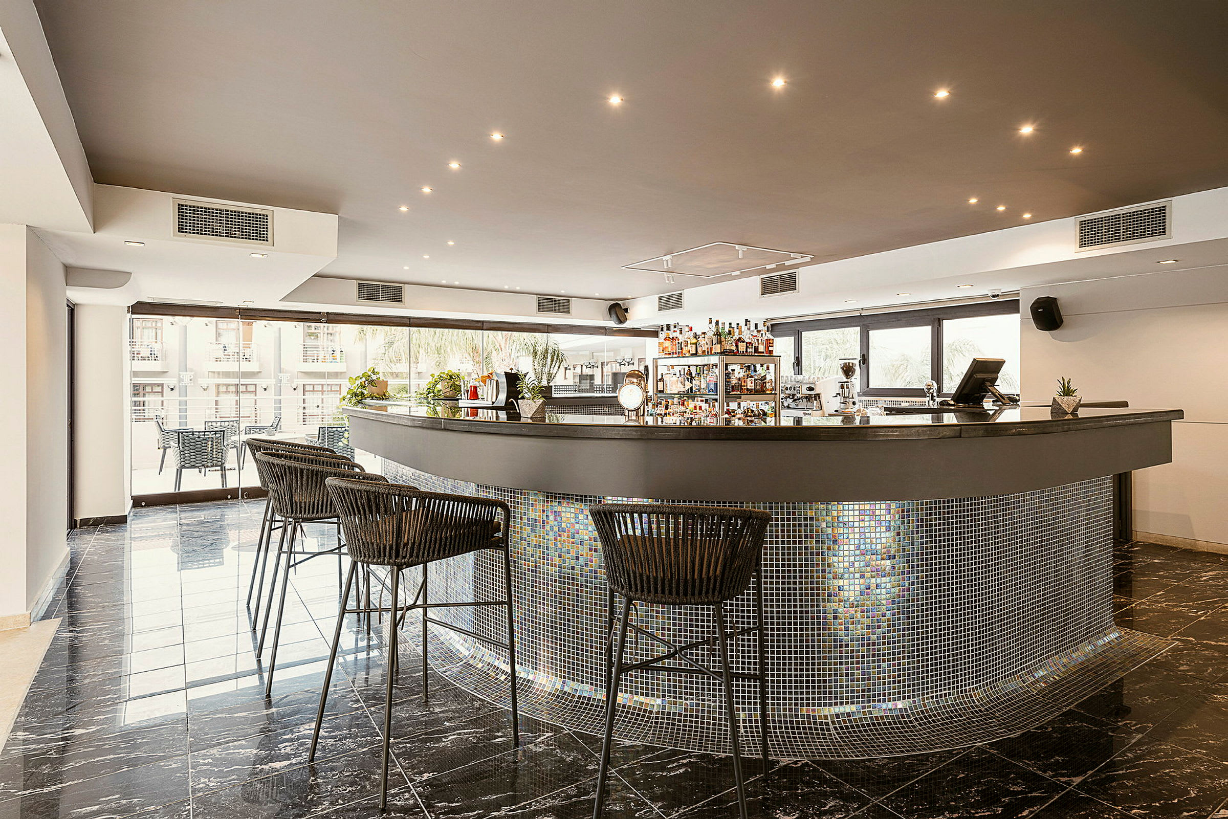 Bild av en modern bar med högstolar framför en bänkskiva med mosaikdetaljer. Fönster i bakgrunden ger naturligt ljus. (AI-genererad text)