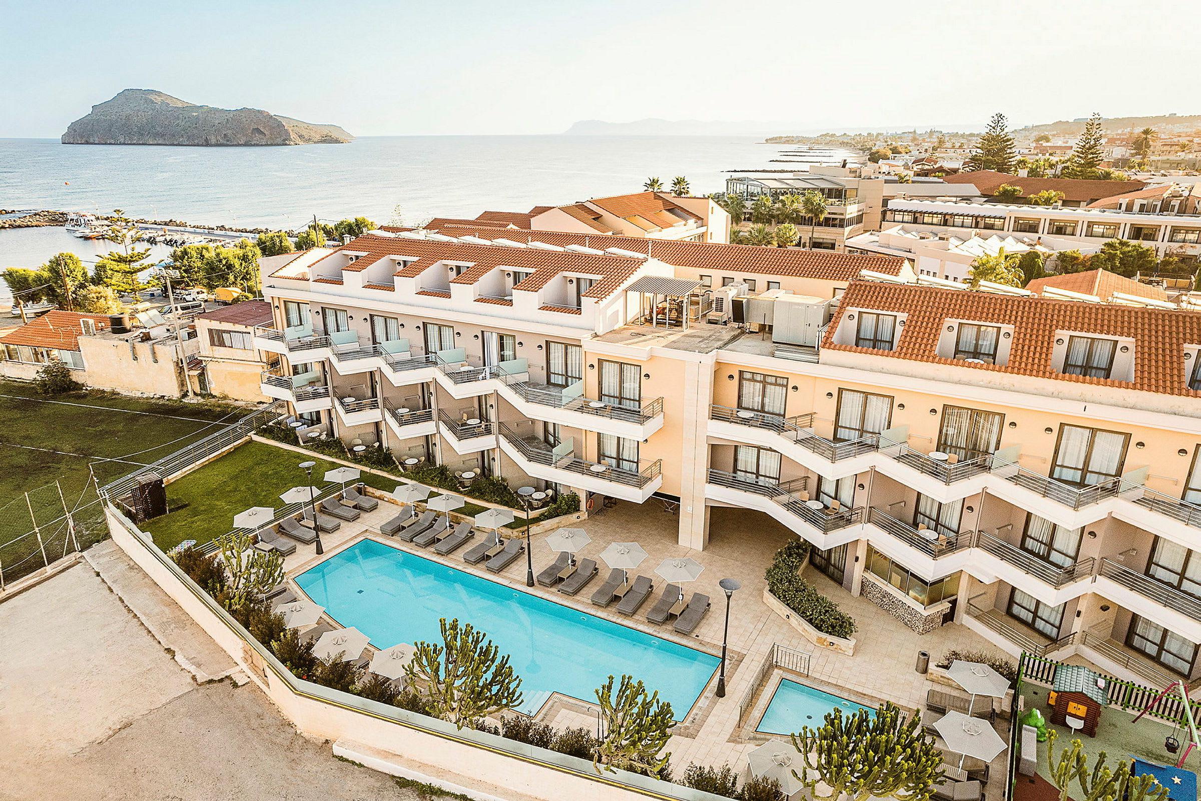 Vy över ett hotellkomplex med en pool, solstolar och parasoller, samt hav och landskap i bakgrunden. (AI-genererad text)