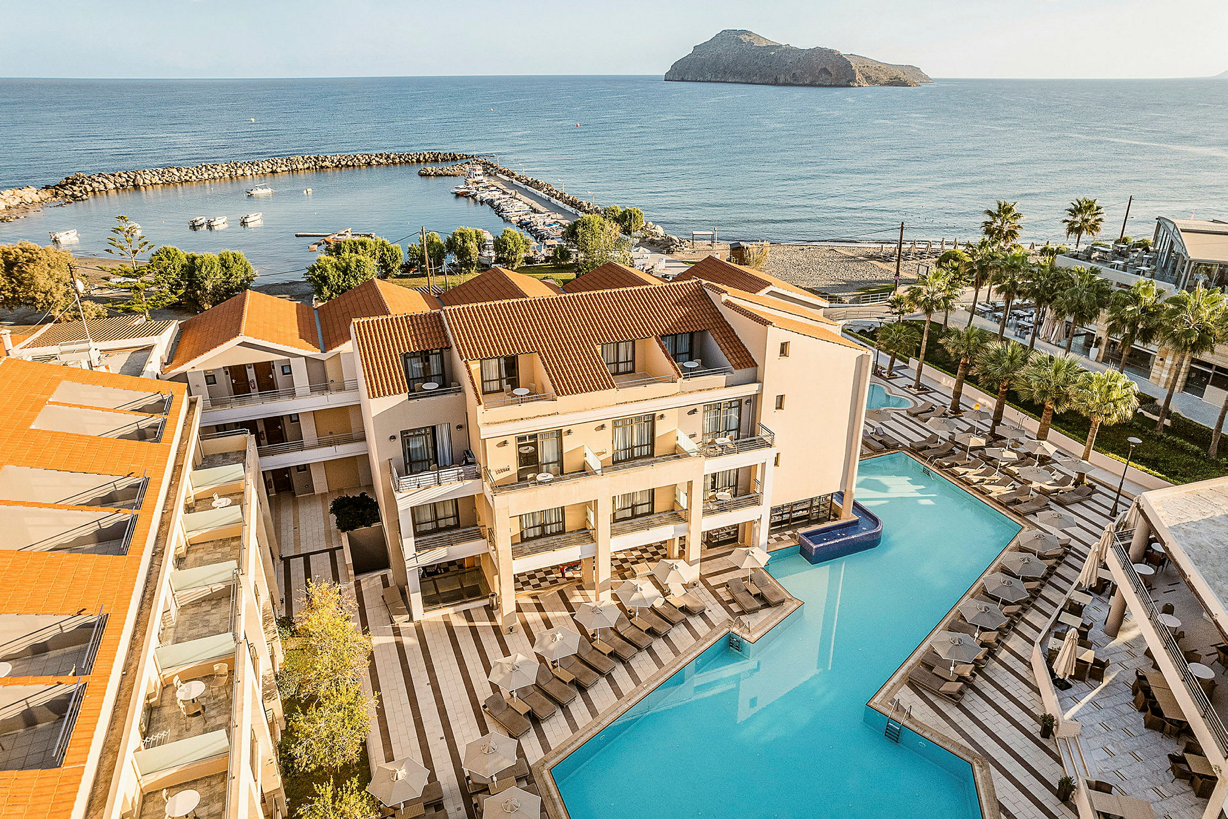 Bild av hotellområde med pool och solstolar intill havet. I bakgrunden syns en liten marina och en ö. (AI-genererad text)