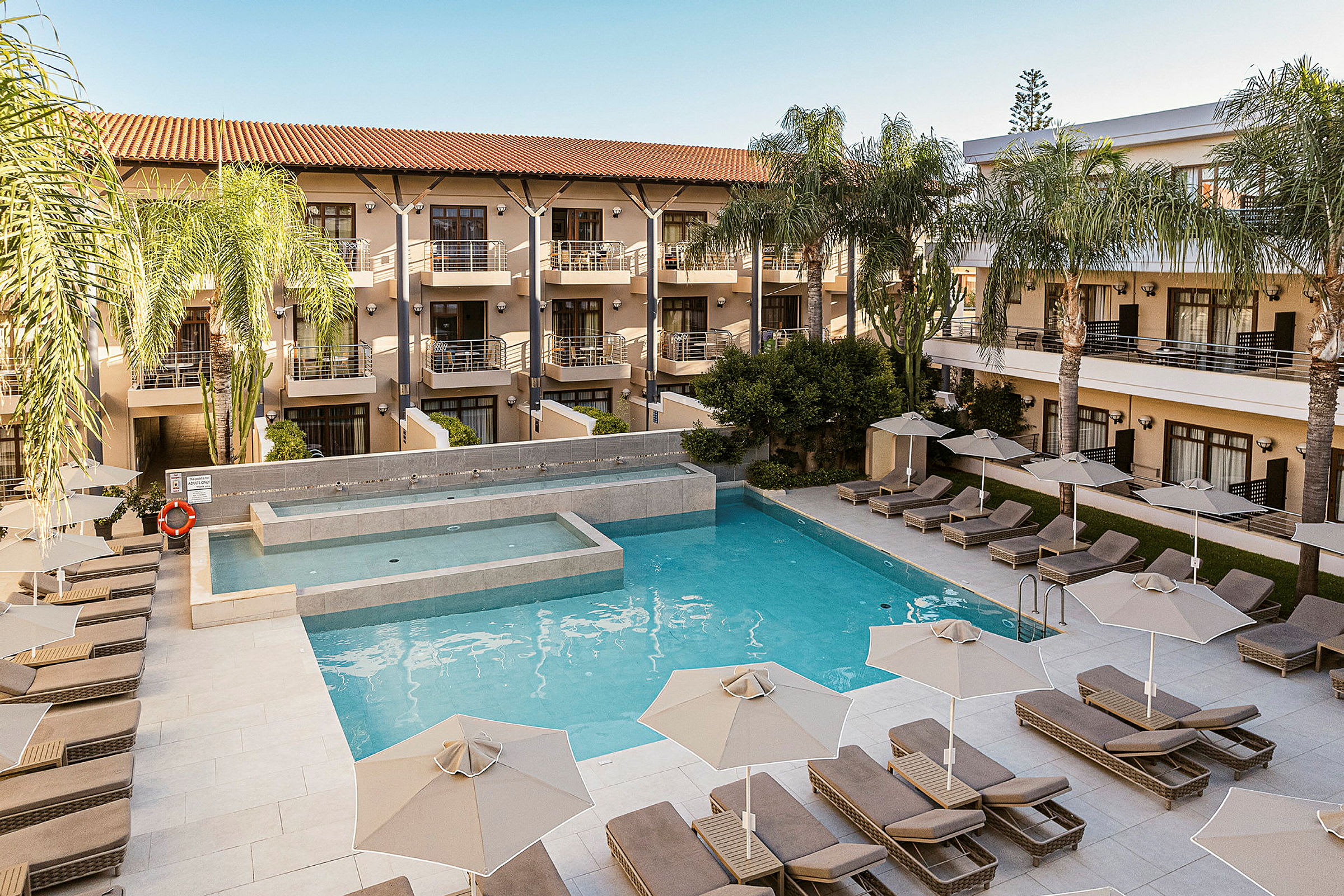 Hotellområde med en pool omgiven av solstolar och parasoller, samt byggnader med balkonger i bakgrunden. (AI-genererad text)