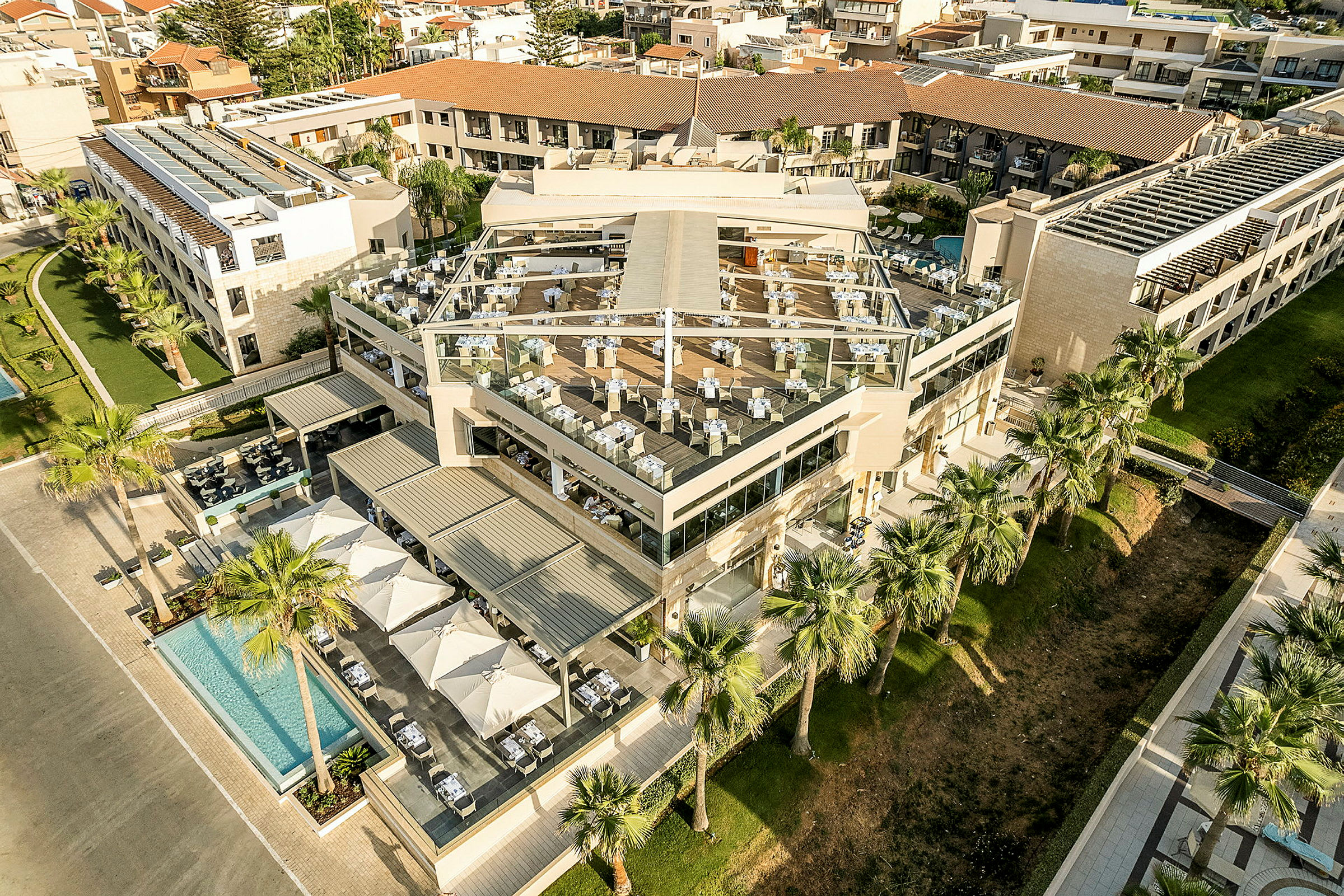 Flygvy över ett modern hotellkomplex med uteservering och poolområde omgivet av palmer. (AI-genererad text)