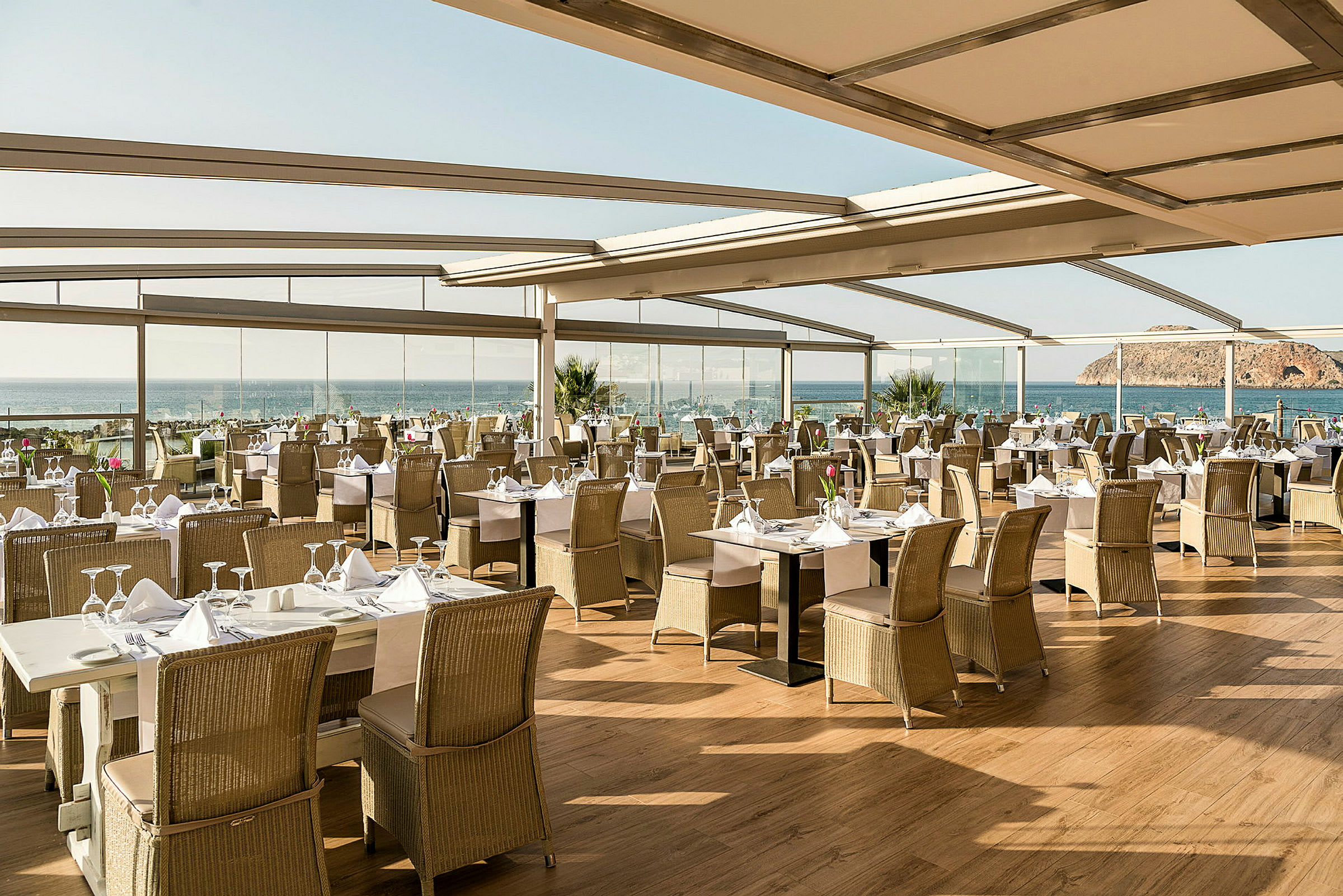En restaurang med dukade bord och utsikt över havet, omgiven av stora glaspartier och tak. (AI-genererad text)