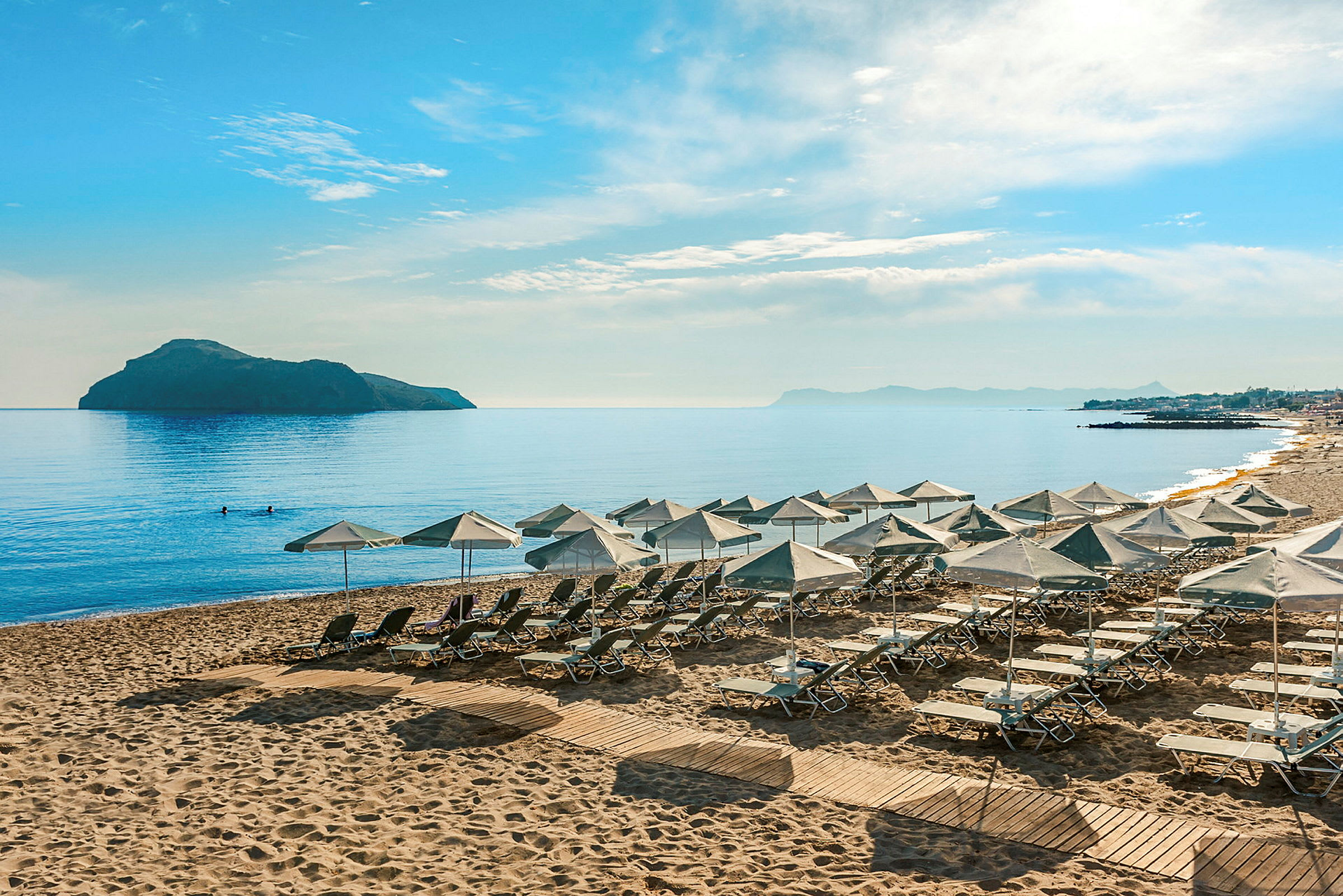 Solstolar och parasoller uppställda på en sandstrand vid en lugn havsutsikt med en ö och berg i bakgrunden. (AI-genererad text)