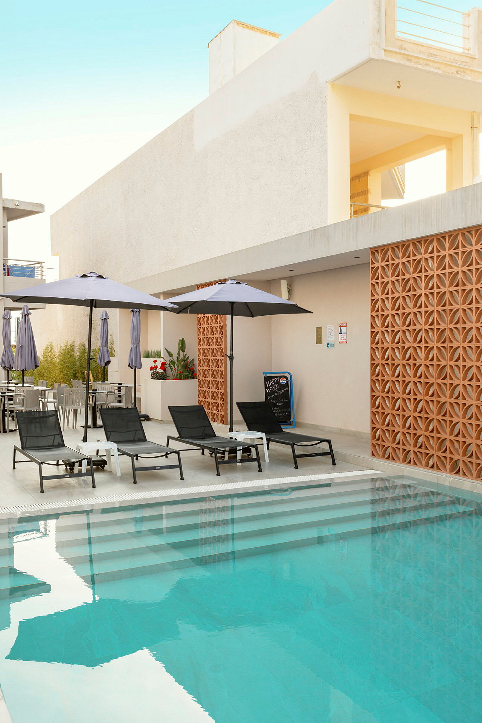 Bild av en pool med klarblått vatten och solstolar med parasoller bredvid. En byggnad med dekorativ vägg och öppningar syns i bakgrunden. (AI-genererad text)