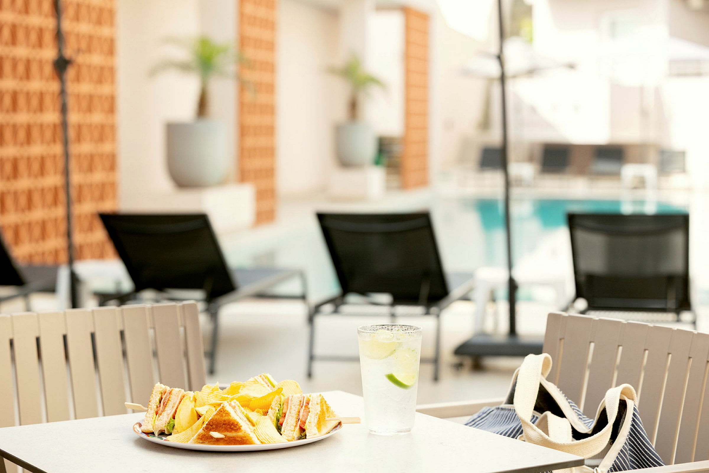 En tallrik med smörgåsar och chips samt ett glas vatten med lime står på ett bord vid en pool med solstolar i bakgrunden. (AI-genererad text)