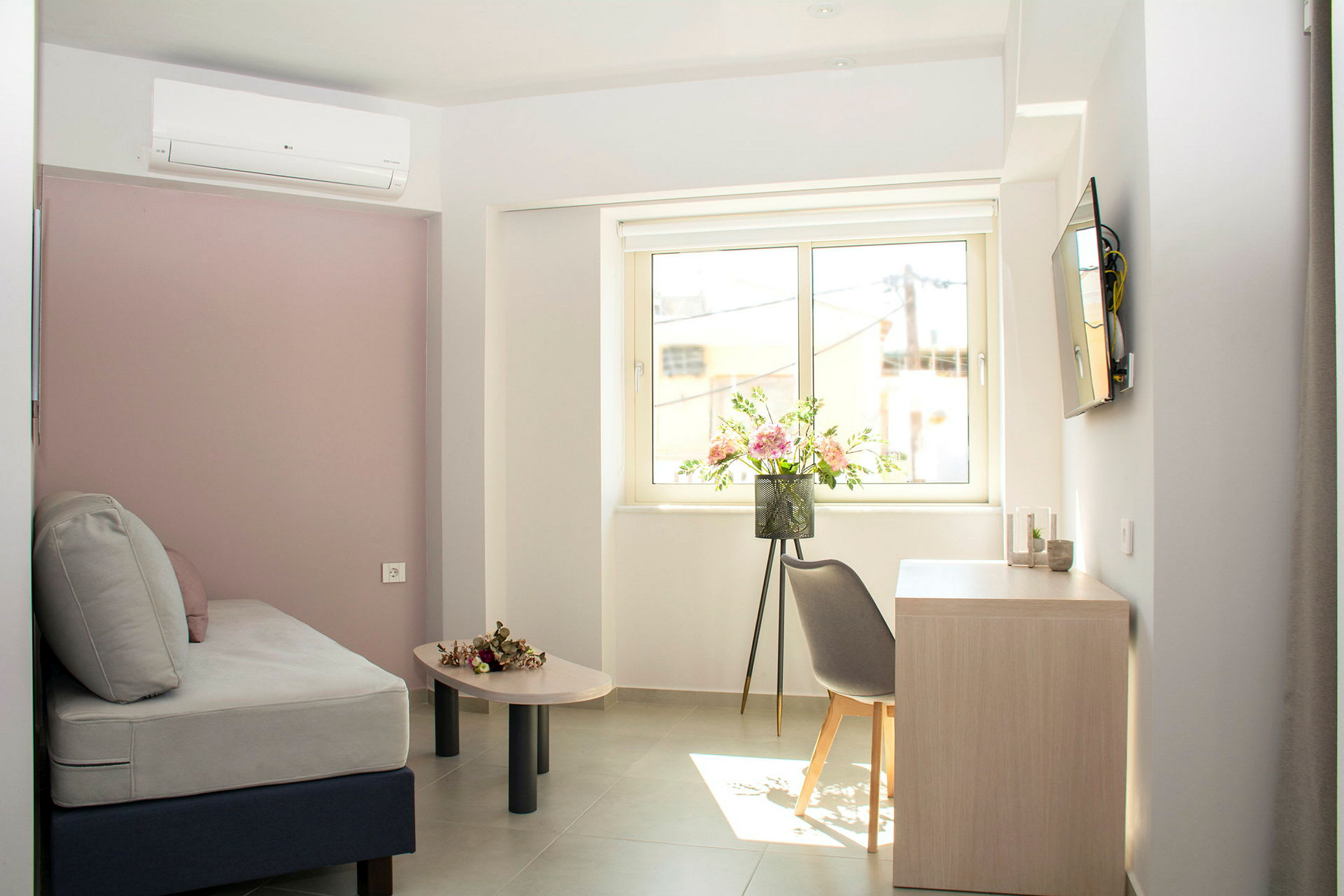Ett ljust hotellrum med grå soffa, blommor på bordet och en fönsterutsikt. (AI-genererad text)