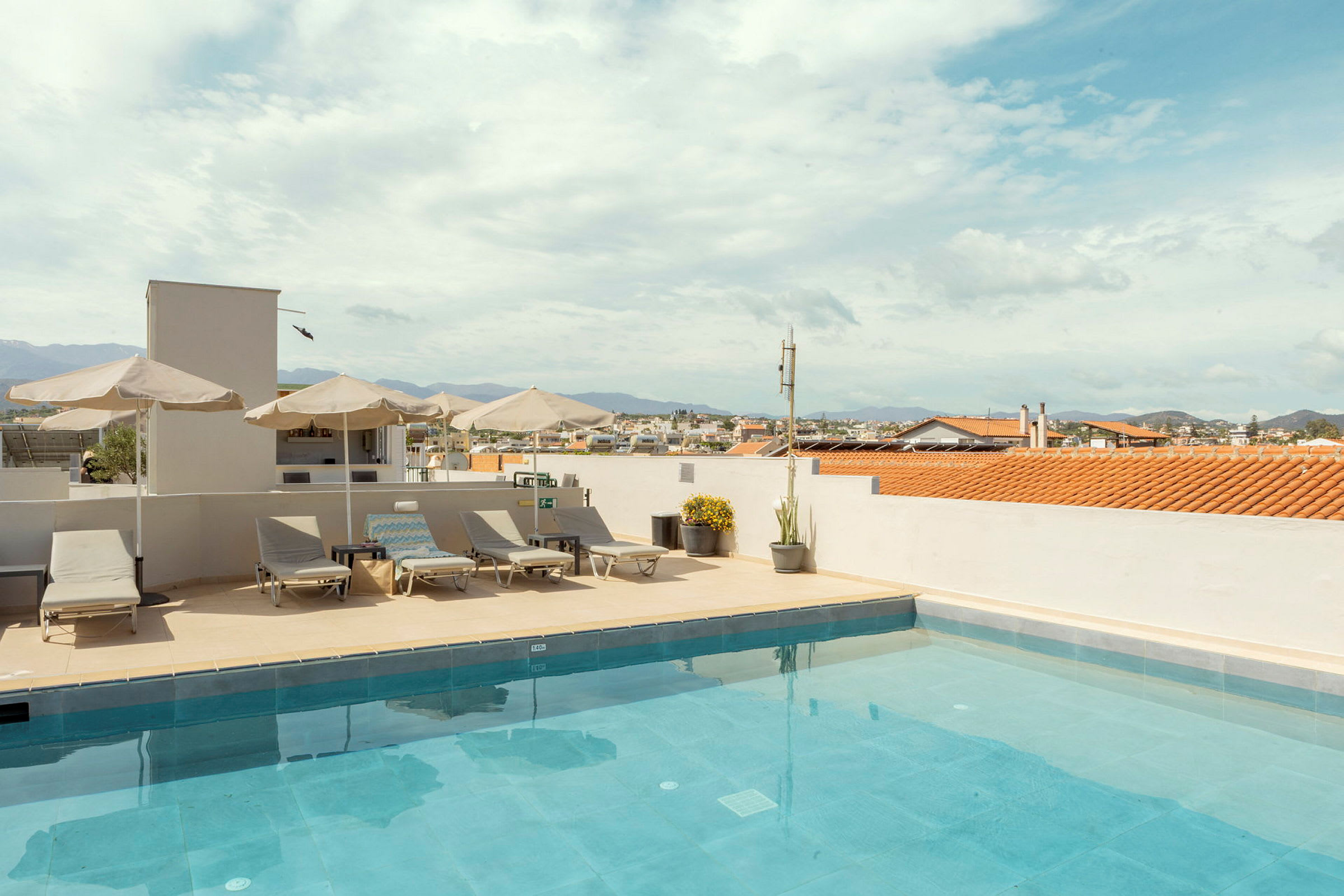 En takterrass med en swimmingpool, solstolar och parasoller under en blå himmel med utsikt över staden. (AI-genererad text)
