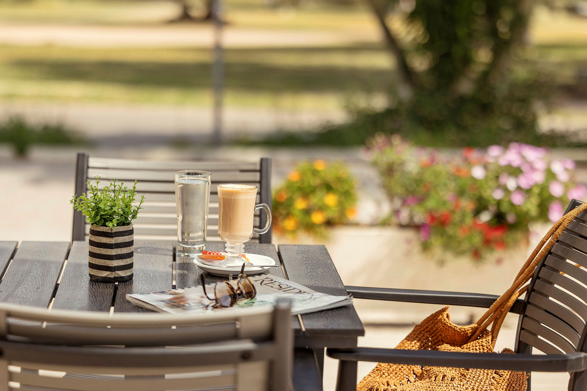 En uteservering med ett bord som har en kaffekopp, ett glas vatten, en tidning och solglasögon. Bakgrunden visar en grönskande miljö och blommor. (AI-genererad text)