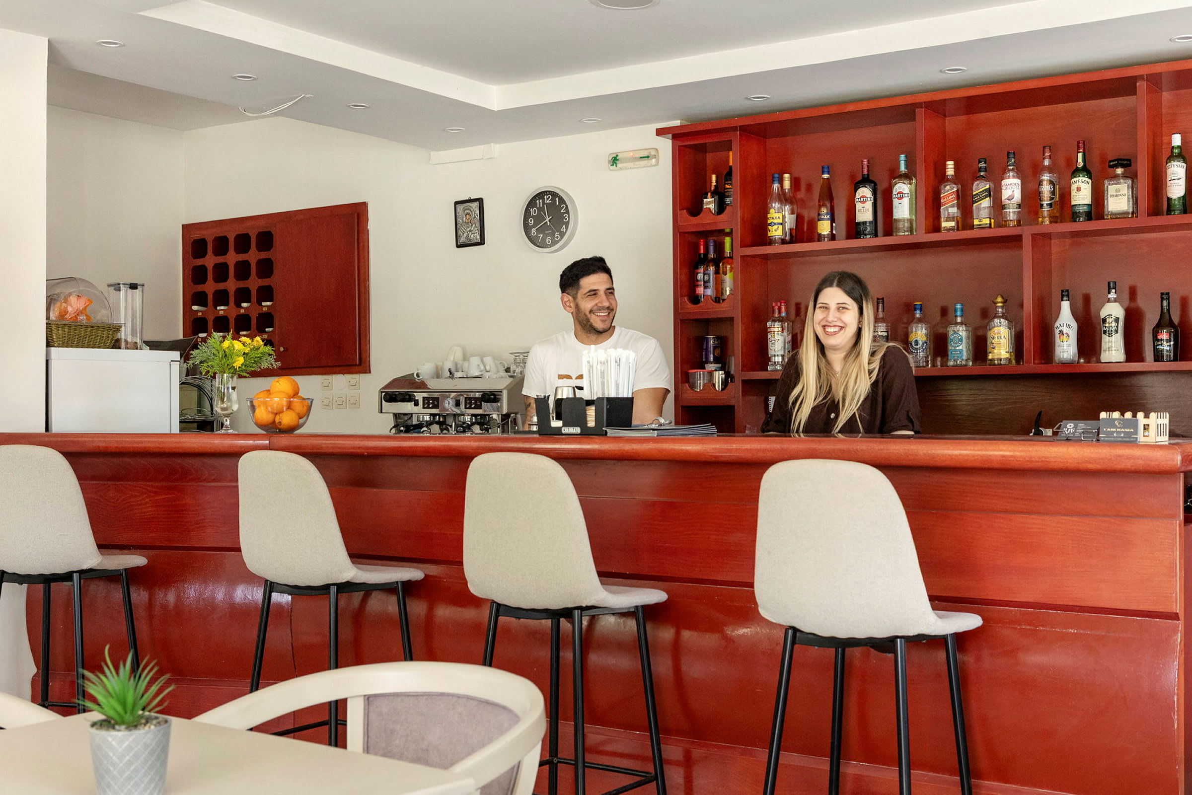 En bar med en trepanel och stolar, flaskor är uppställda på hyllor, och två personer står bakom baren. (AI-genererad text)