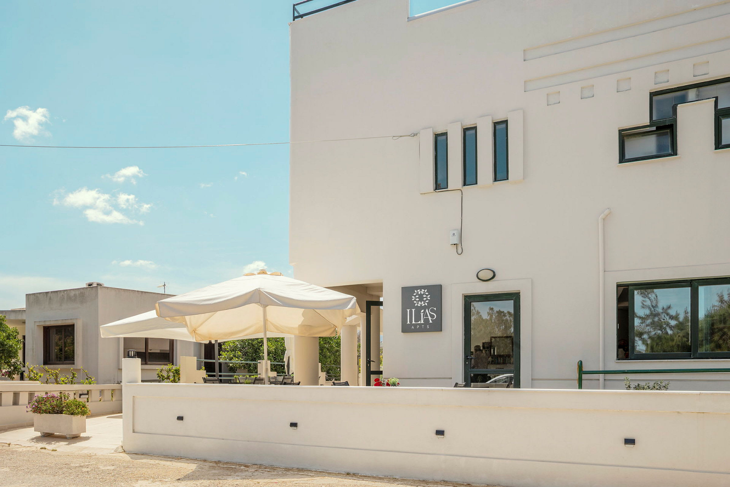 Bild av en vit byggnad med flera fönster och en skylt som säger 'Ilias Apts'. Framför byggnaden finns en uteplats med ett stort parasoll och blomsterlådor. (AI-genererad text)