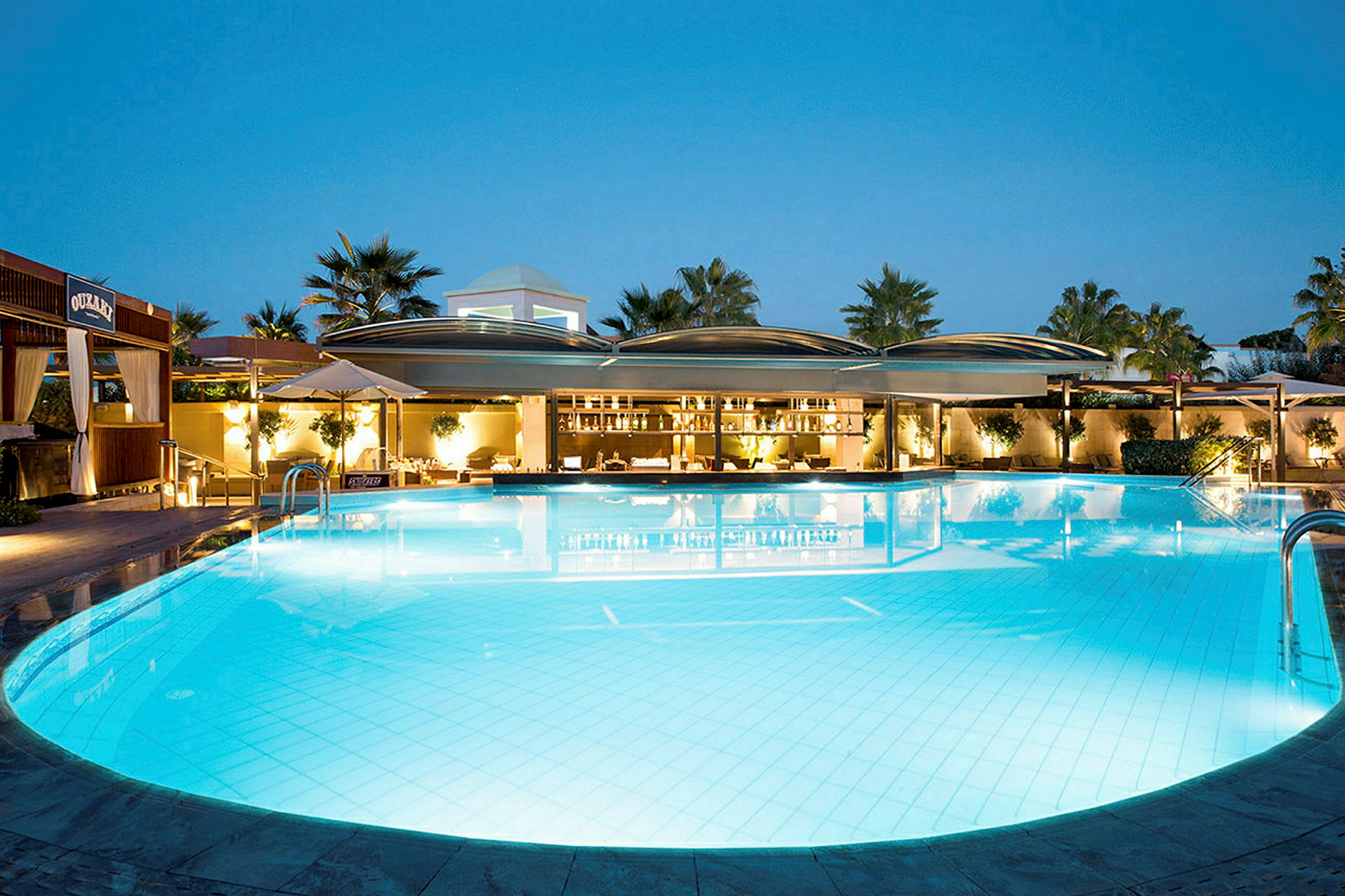 En upplyst pool vid en resort under blå kvällshimmel, omgiven av växtlighet och byggnader. (AI-genererad text)