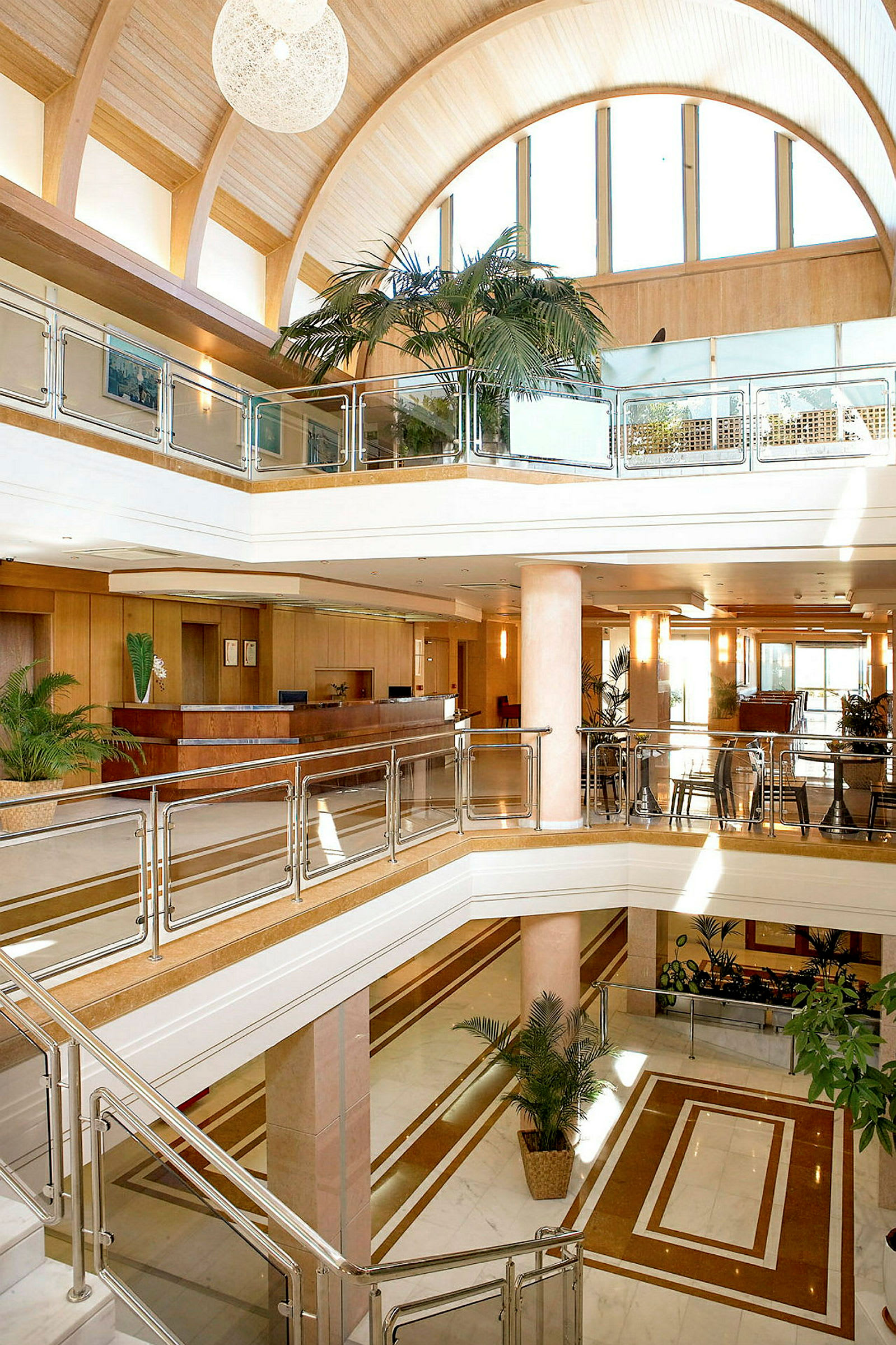 Interiören i en modern hotellobby med flera våningar. Glasräcken, trädetaljer och stora palmer ses tillsammans med en reception i bakgrunden. (AI-genererad text)
