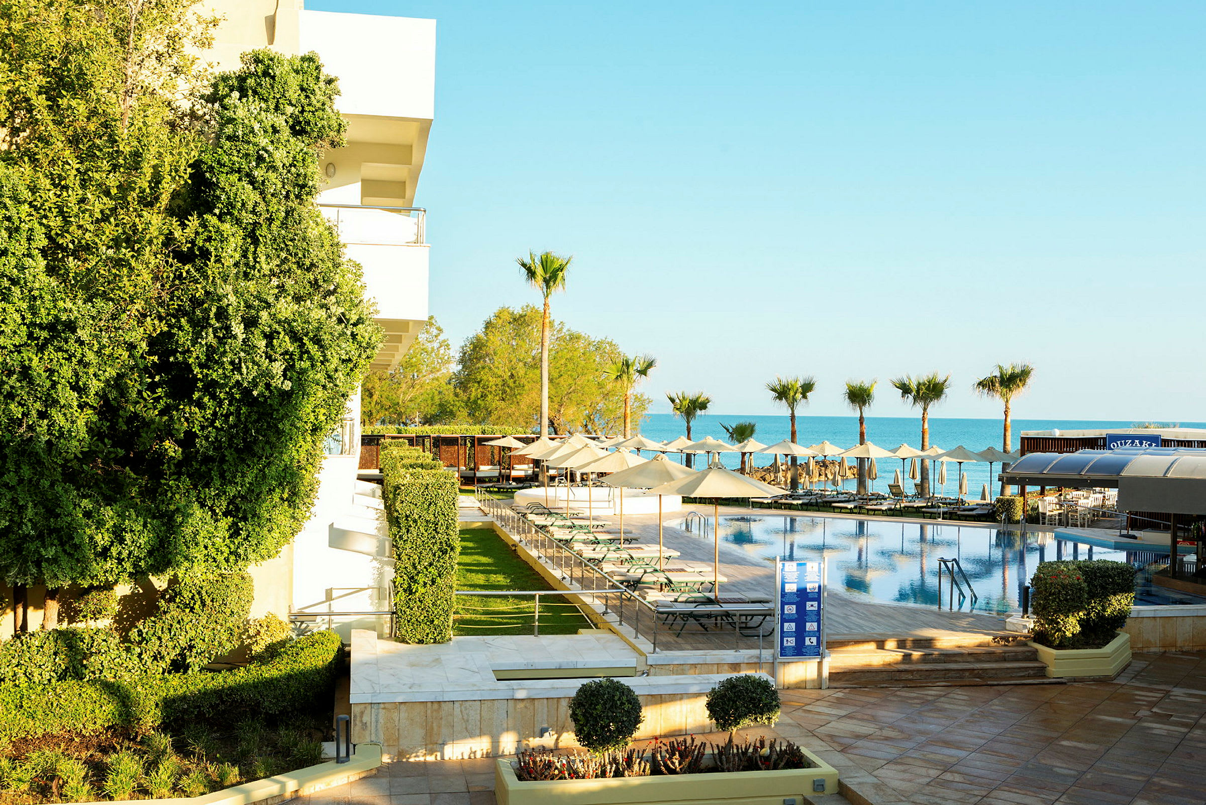 Bild som visar ett hotellområde med en swimmingpool och solstolar, omgivna av palmer och grönska nära havet. (AI-genererad text)