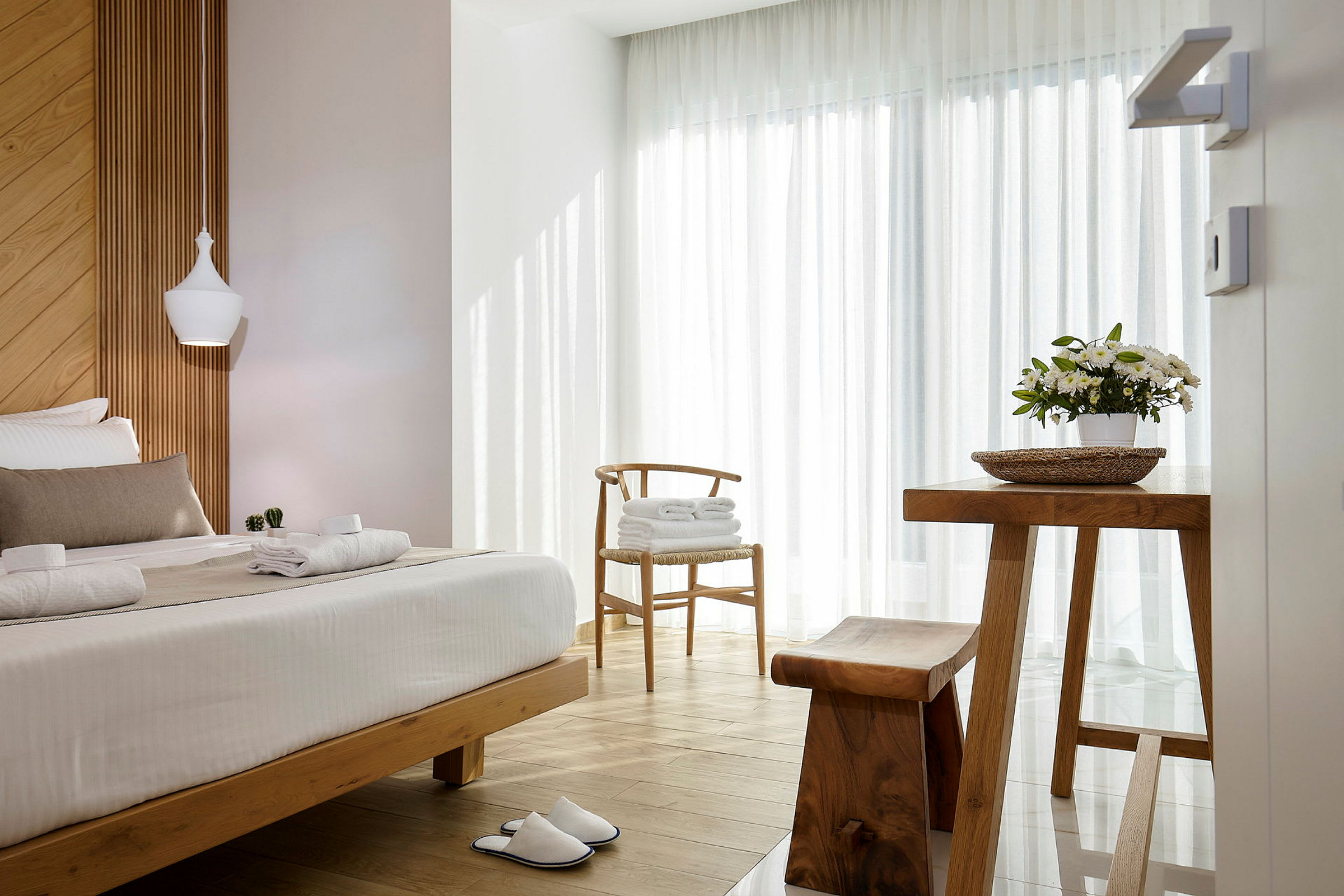 Ett ljust och luftigt hotellrum med trädetaljer, en bäddad säng, en blomkruka på ett bord och ett par vita tofflor på golvet. (AI-genererad text)