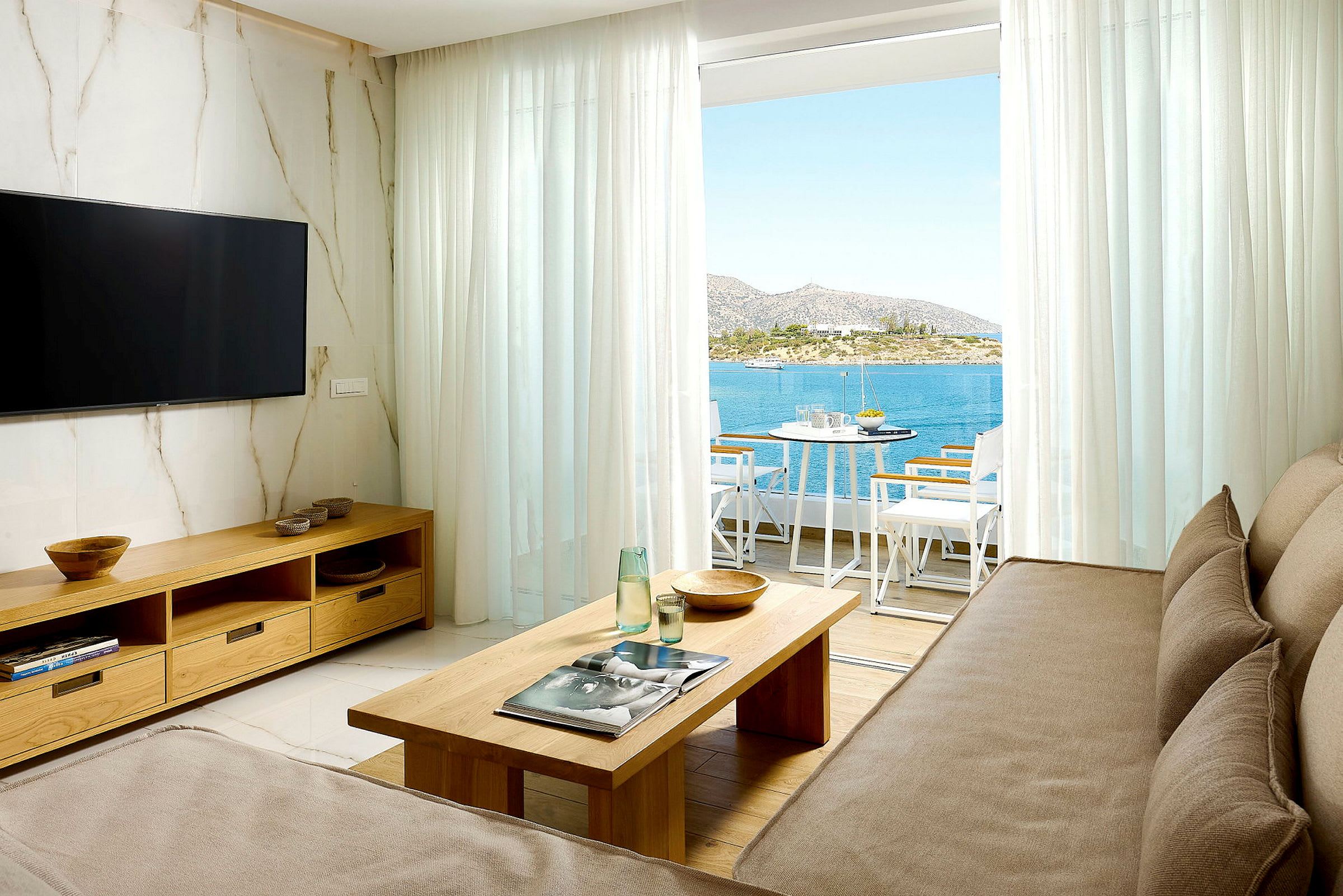 Ett vardagsrum med soffa, soffbord, TV och utsikt över havet genom en balkong med sittmöbler. (AI-genererad text)