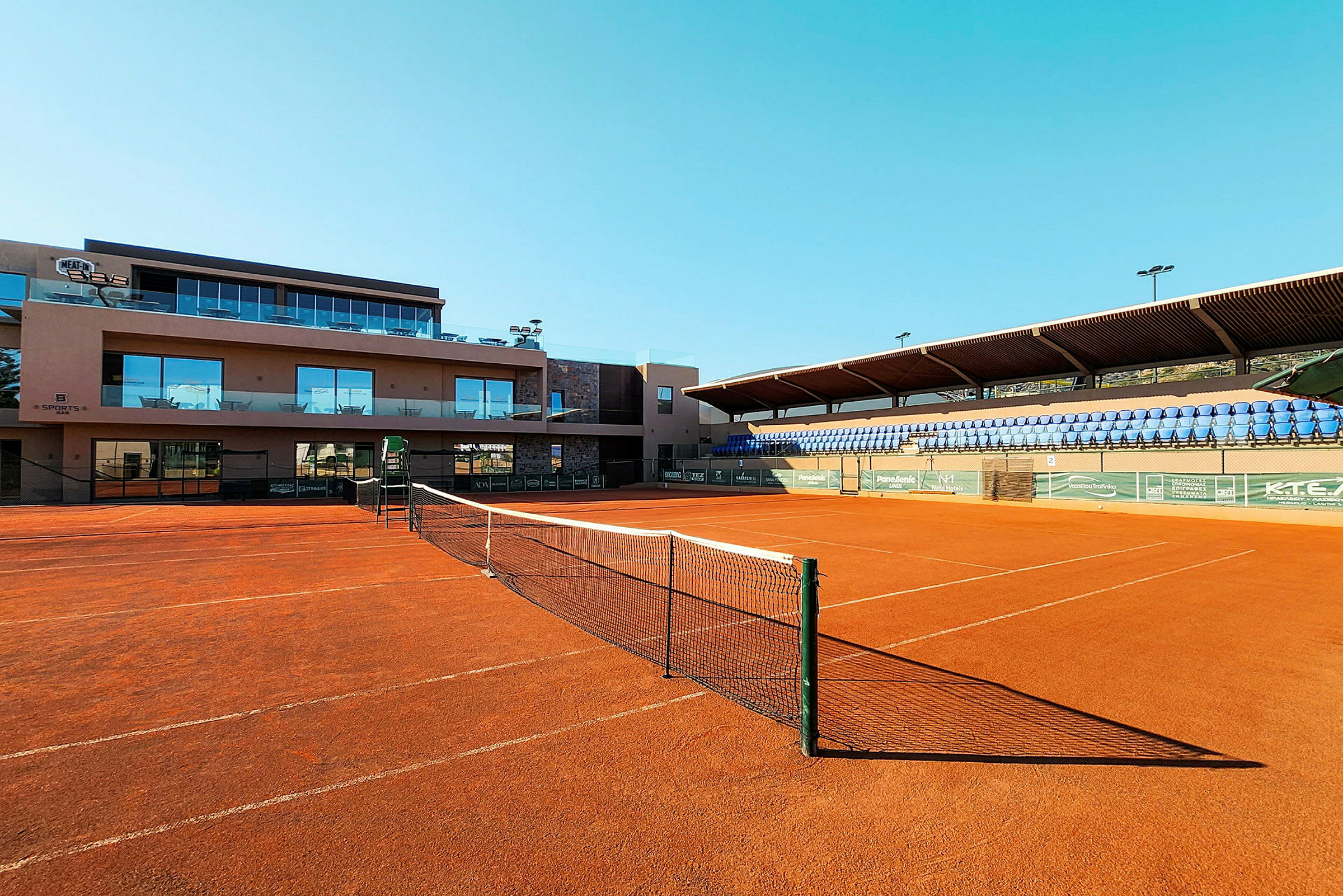 En tennisbana med rödgrus och nät framför en byggnad och läktare. (AI-genererad text)