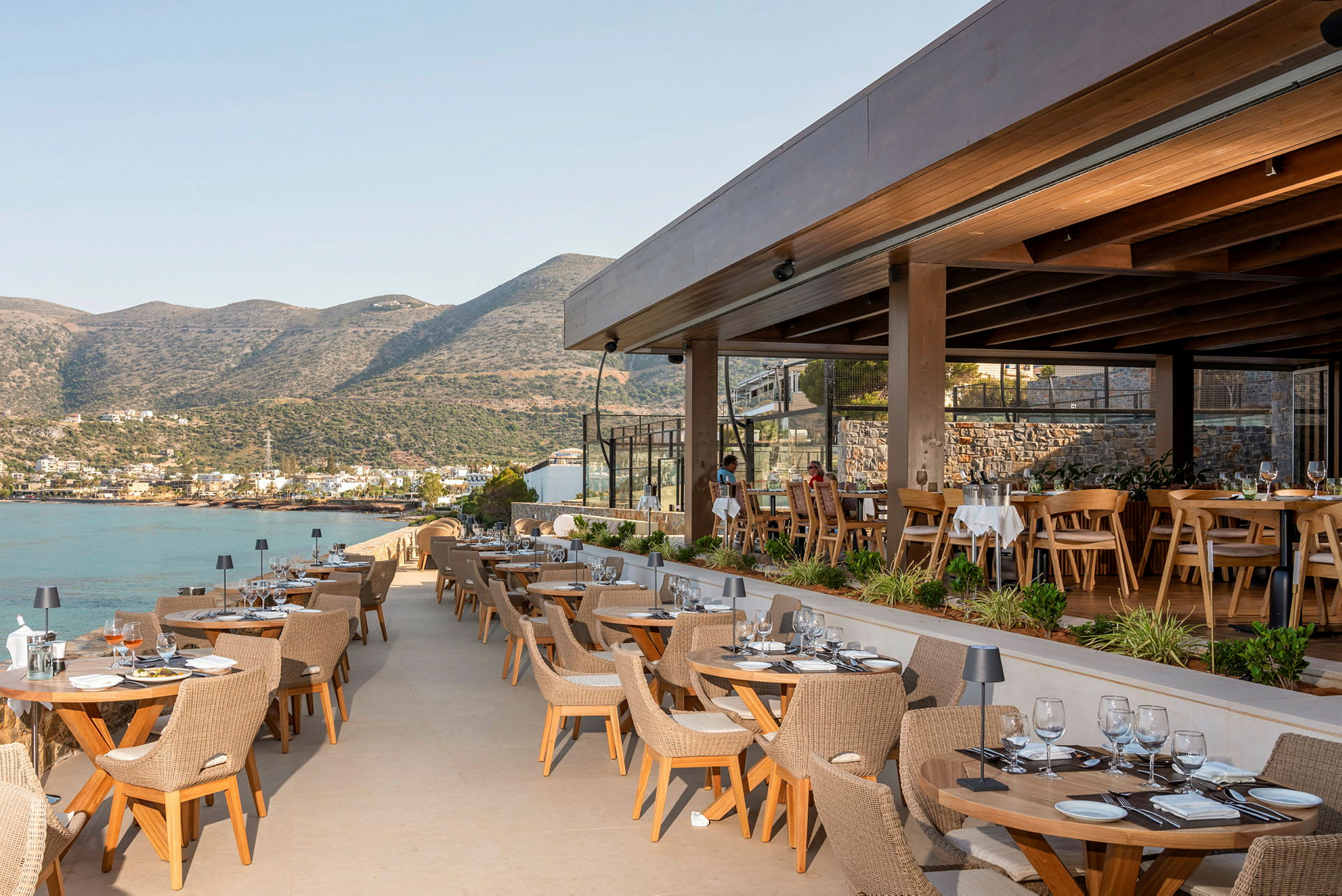 Bild av en utomhusrestaurang med utsikt över en havsvik och berg i bakgrunden. (AI-genererad text)