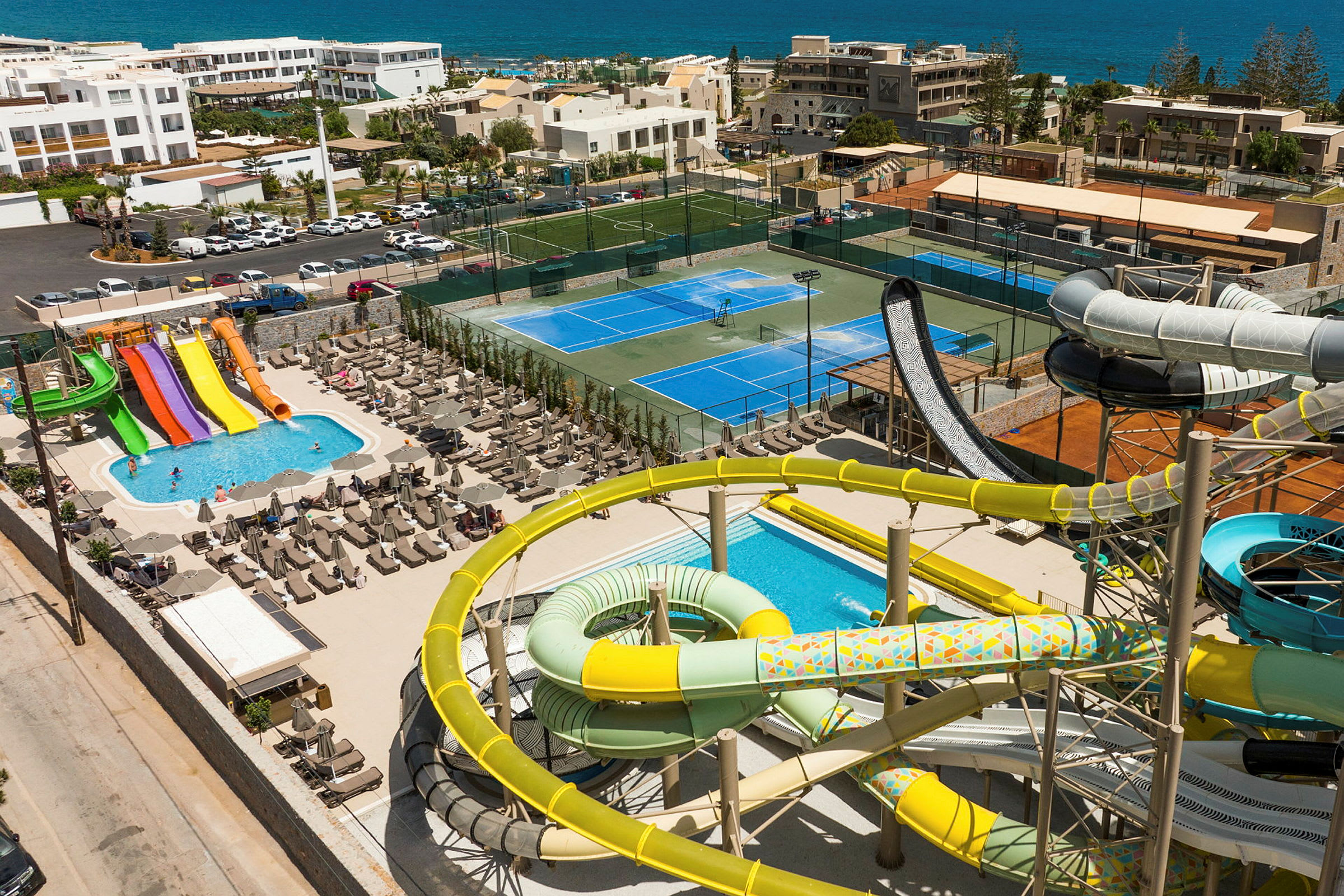 Bild av en ferieanläggning med vattenrutschbanor, pool, tennisbanor och vy över havet. (AI-genererad text)