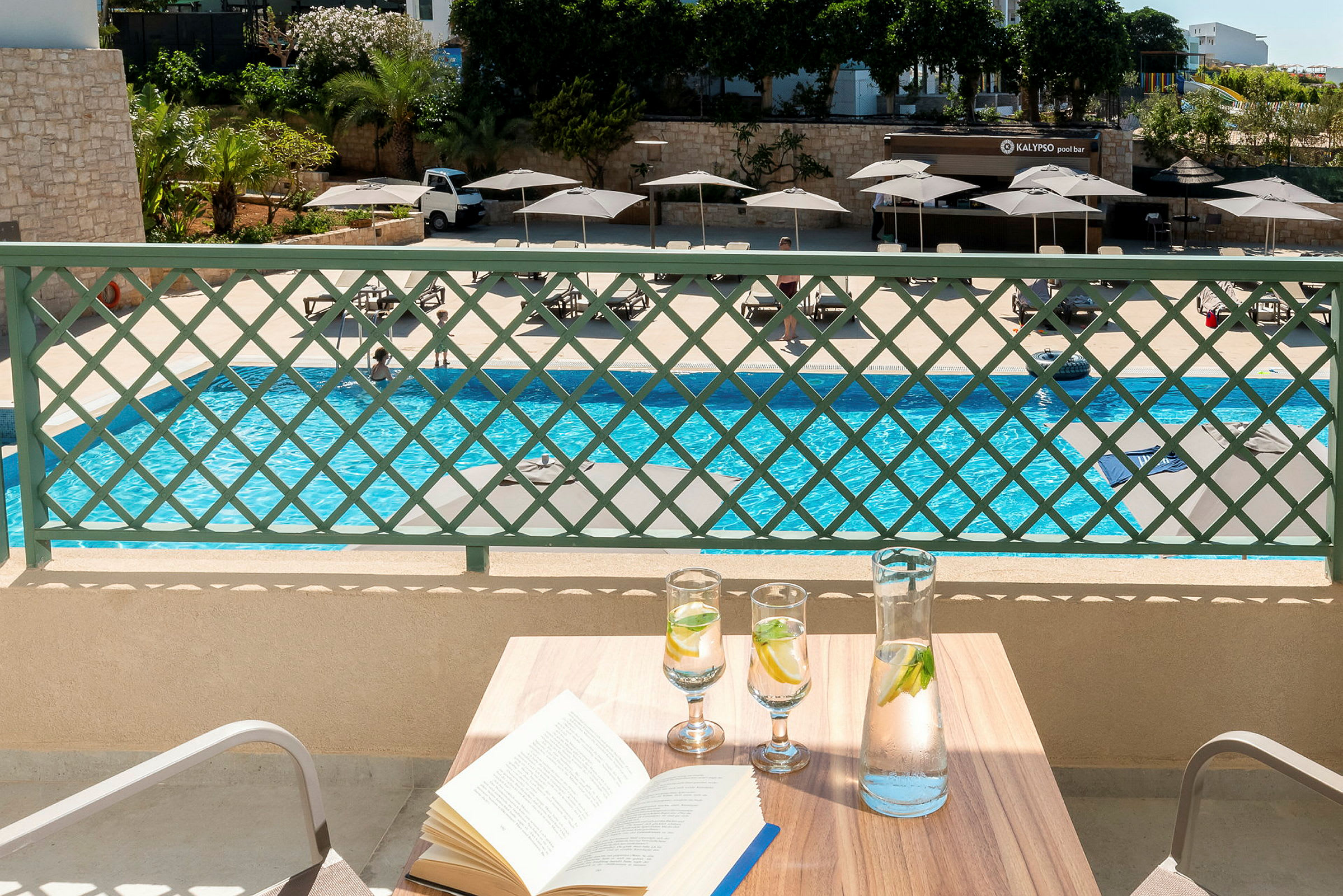 Bild av ett bord med en bok och glas vatten, med en pool och solstolar i bakgrunden. (AI-genererad text)