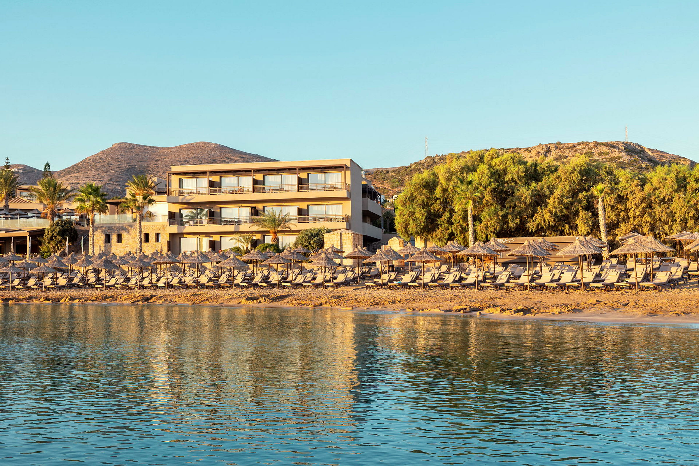 En semesteranläggning vid stranden med solstolar och parasoller i förgrunden och ett hotell bakom. (AI-genererad text)