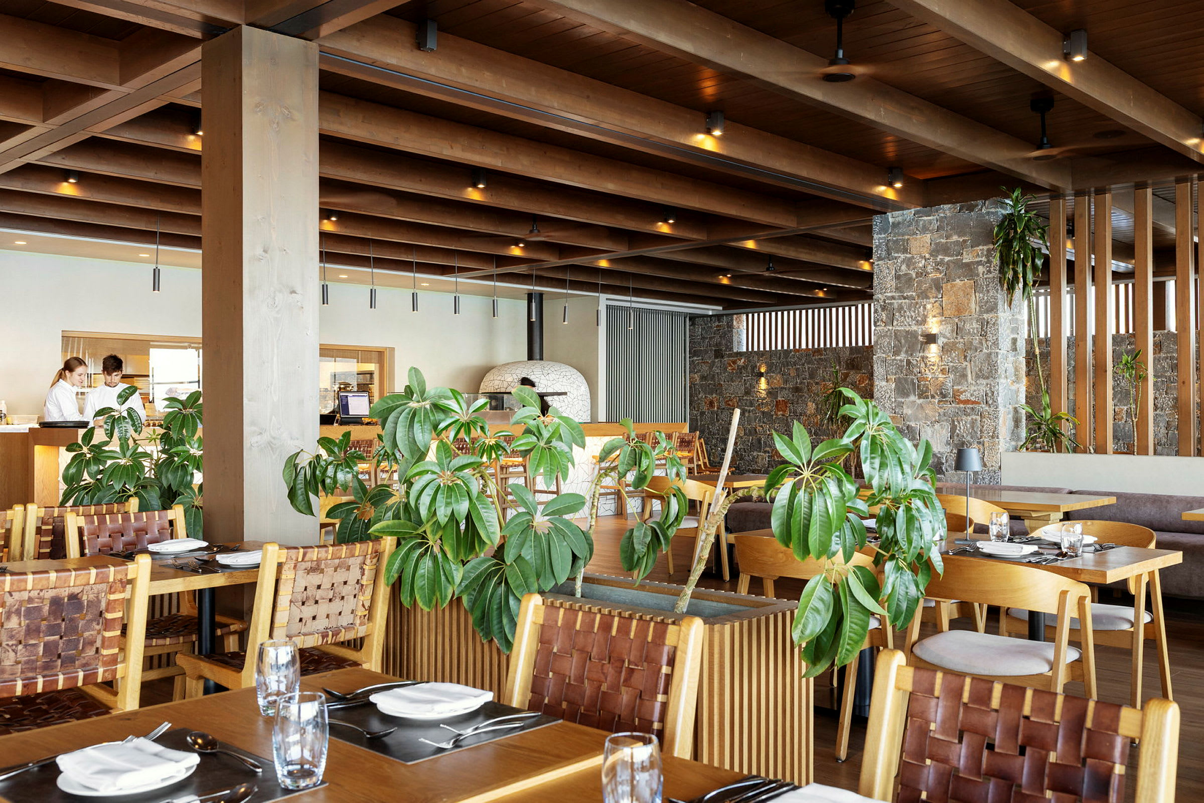 Ett modernt restauranginteriör med trämöbler, gröna växter och stenväggar. (AI-genererad text)