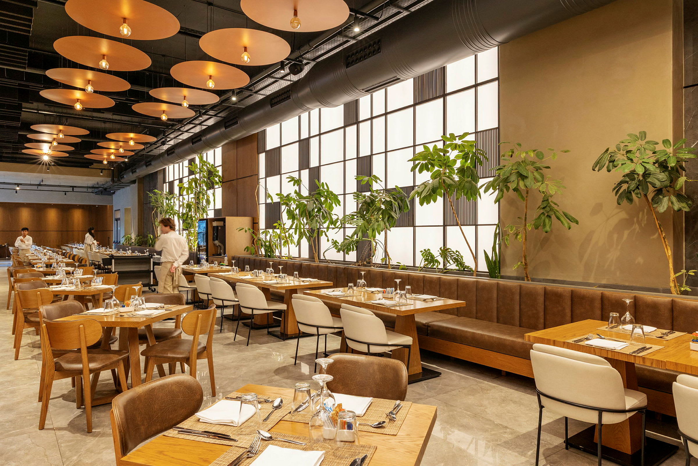 En modern restaurang med träbord och stolar, gröna växter längs en vägg och en servitör synlig i bakgrunden. (AI-genererad text)