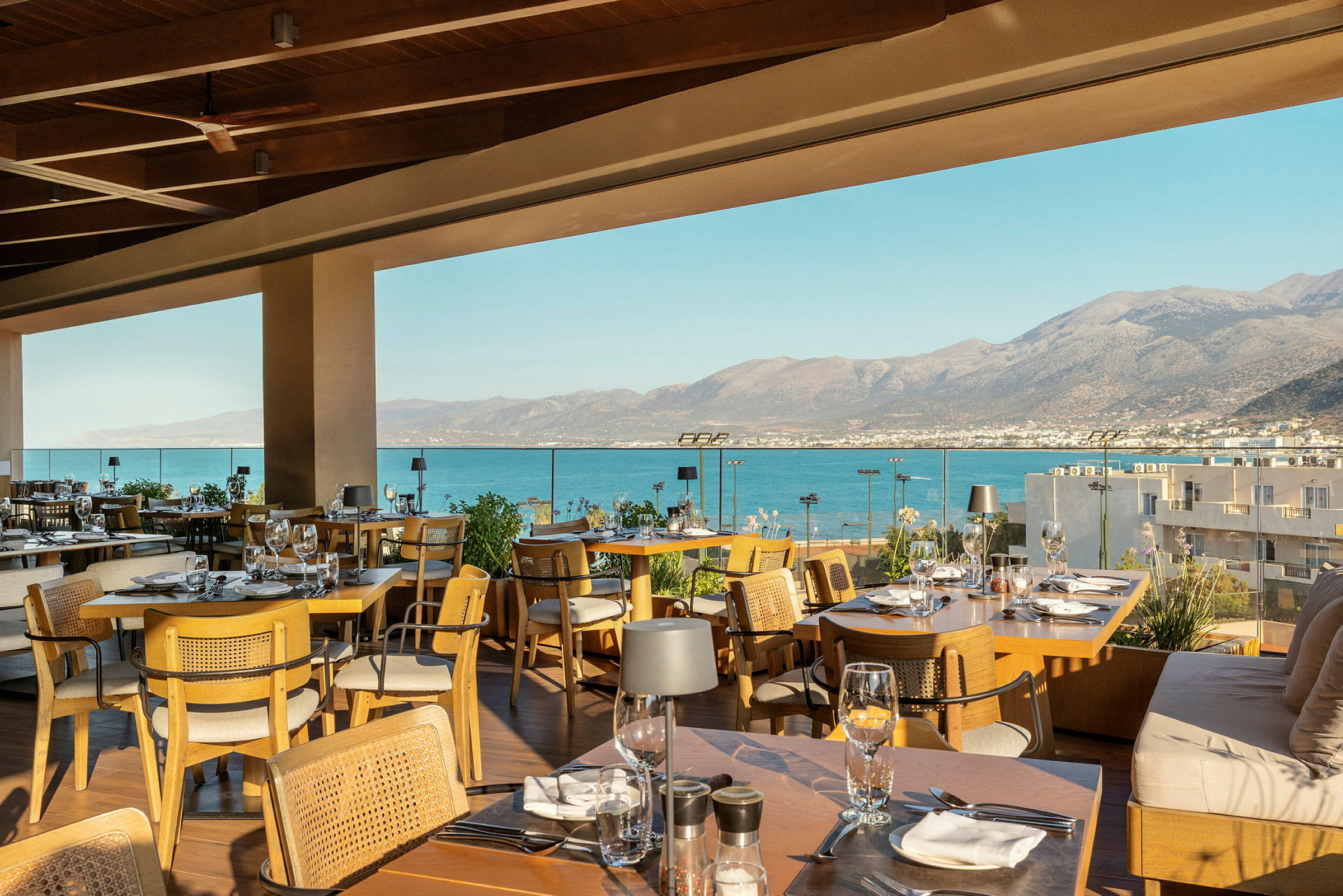 En restaurang med utsikt över havet från en terrass. Uteplatsen är möblerad med träbord och stolar, och bergen i bakgrunden sträcker sig bortom vattnet. (AI-genererad text)