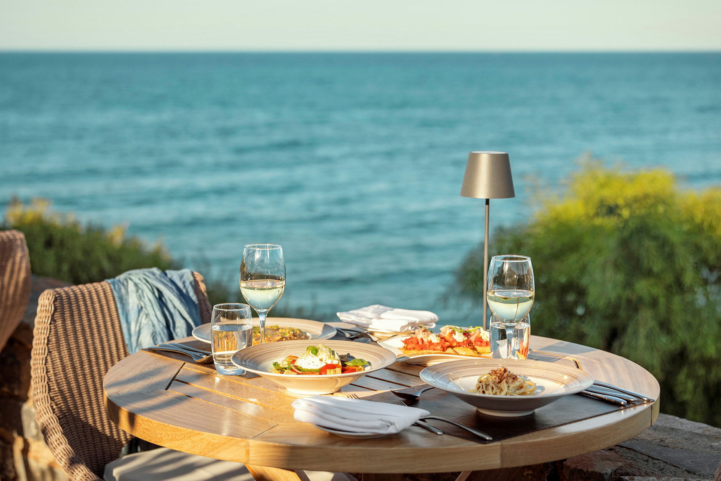 En utomhusservering vid havet med en rund bord fylld med tallrikar mat, vinglas och bestick. (AI-genererad text)