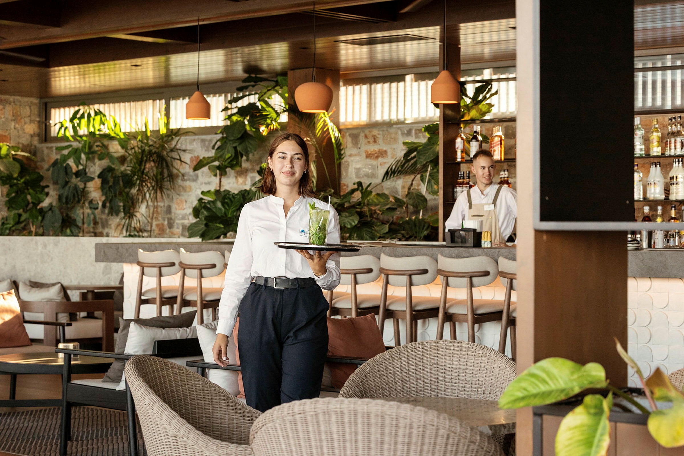 En servitris bär en bricka med en drink i en trevlig lounge med växter och en bardisk i bakgrunden. (AI-genererad text)