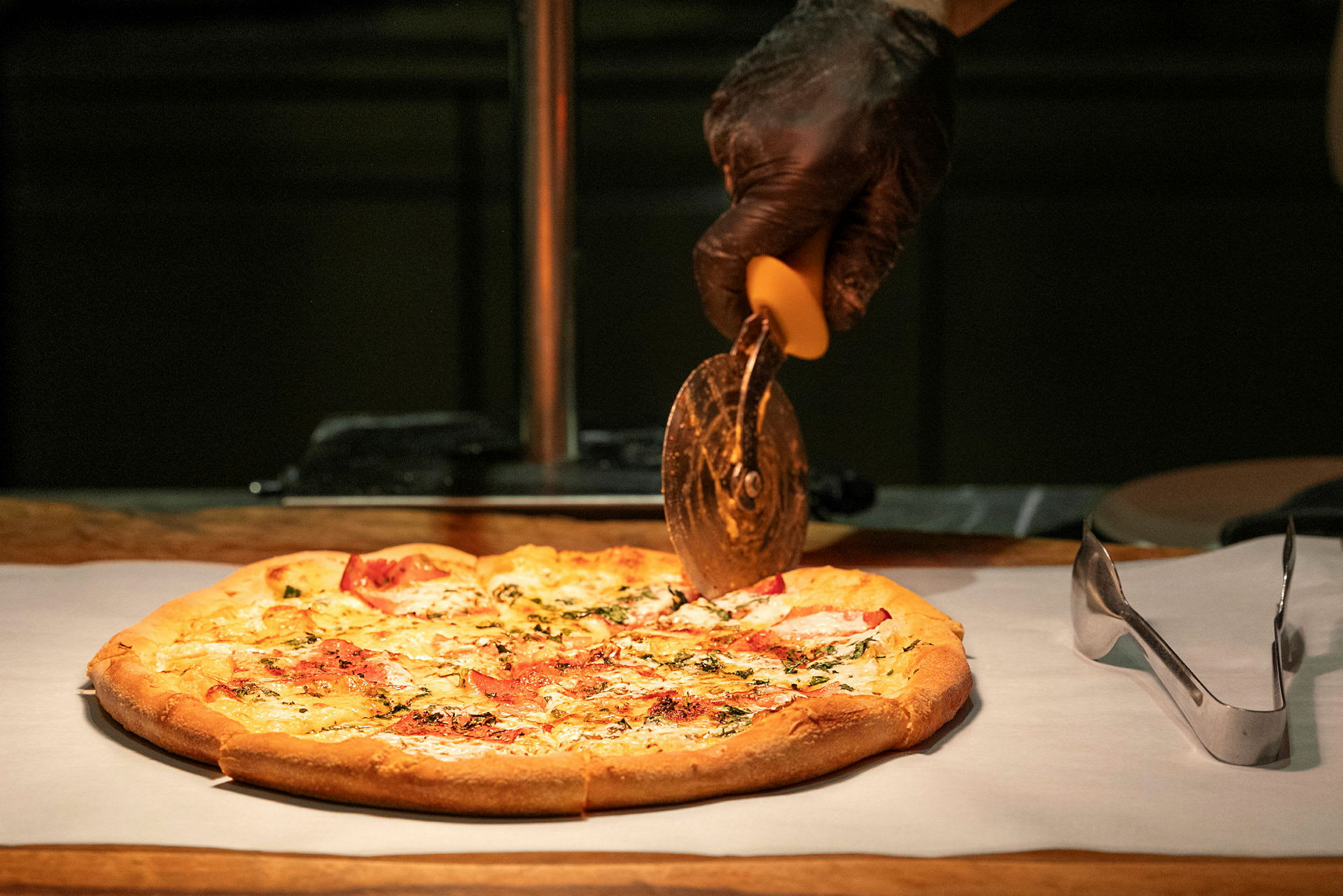 En pizza på ett bakplåtspapper skärs med en pizzaskärare som hålls av en hand i svart handske. (AI-genererad text)