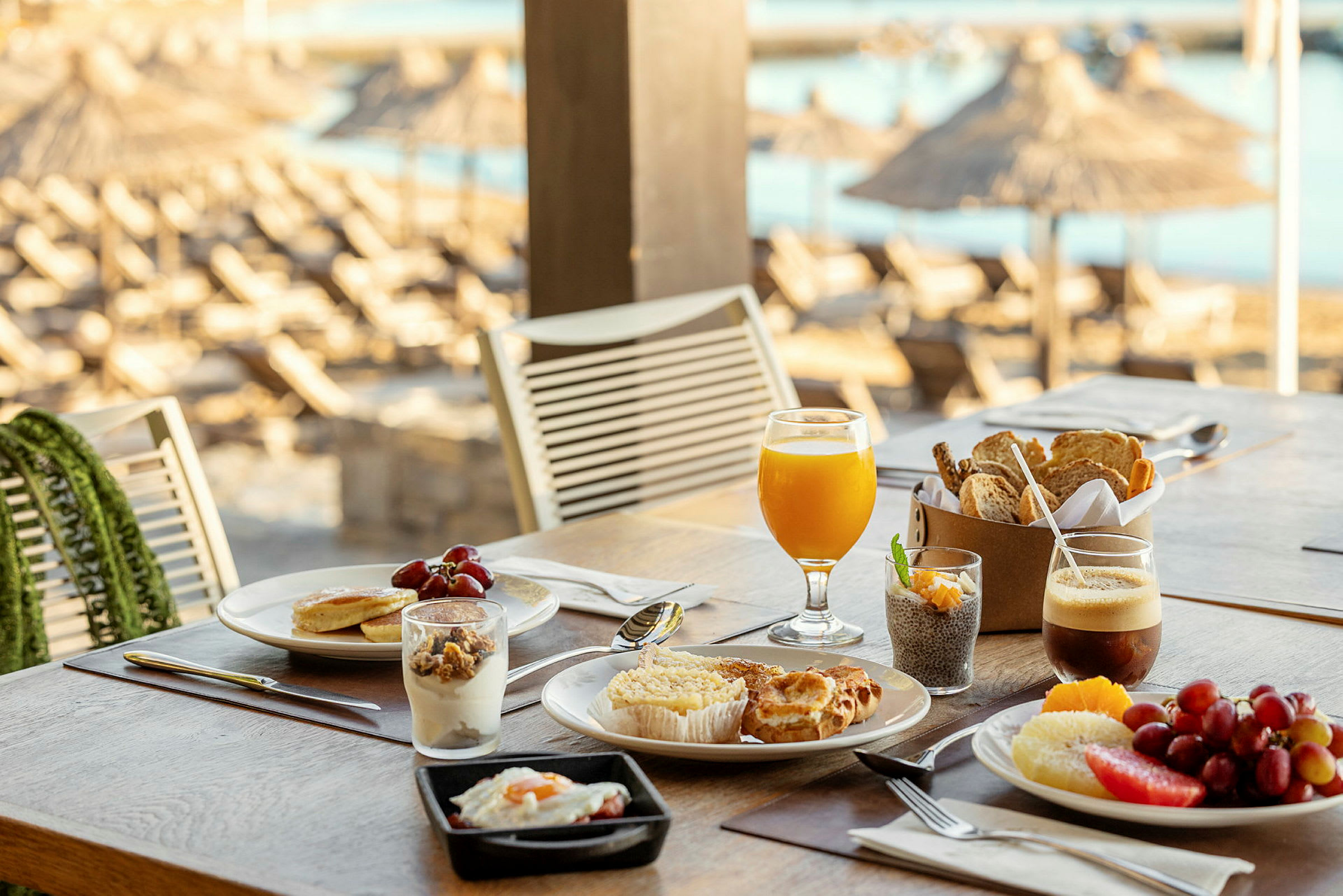 Ett bord med en frukostuppsättning, inklusive glas med juice och kaffe, bakverk, frukt och yoghurt. I bakgrunden syns parasoller på stranden. (AI-genererad text)